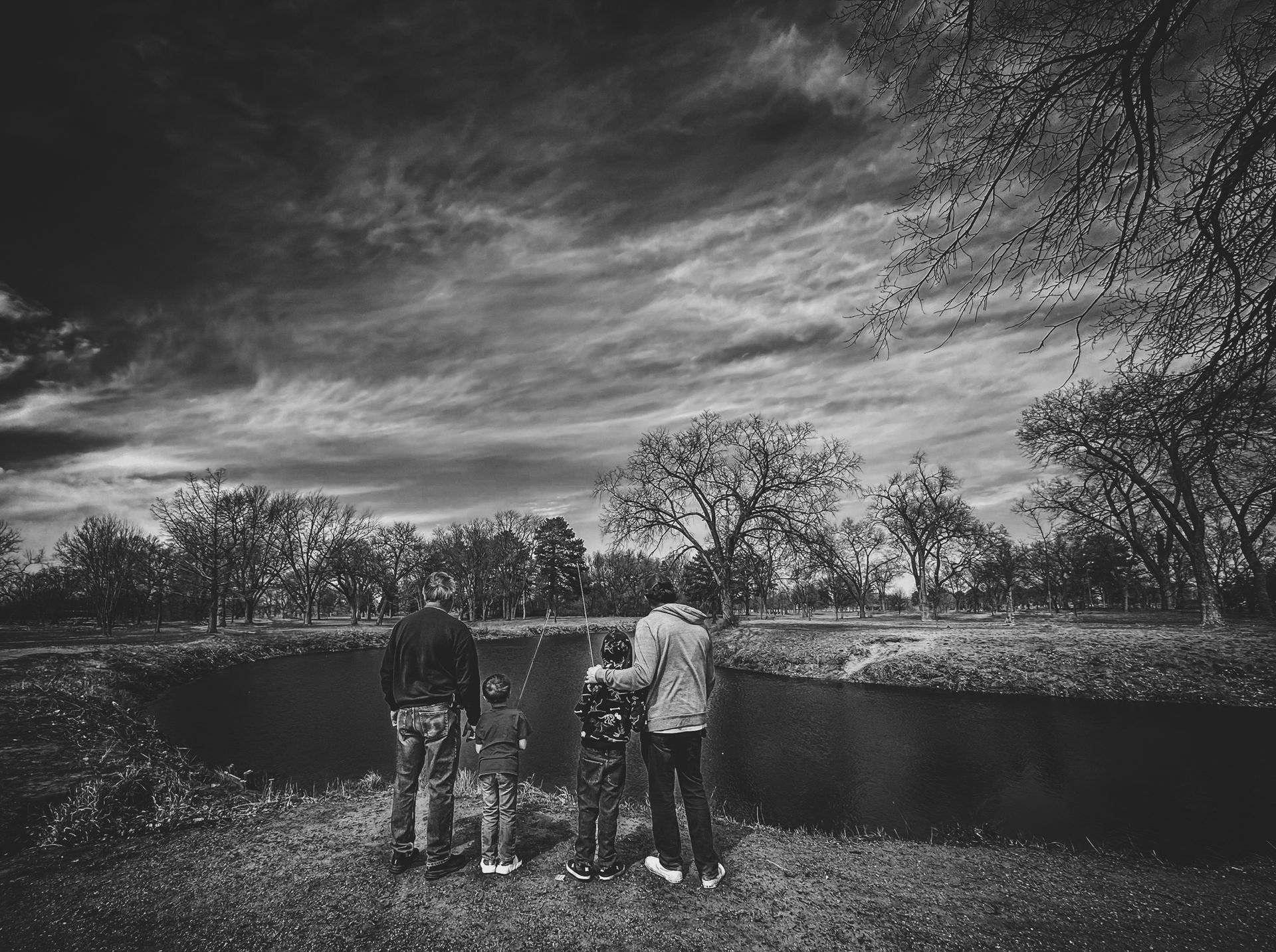 Family of four standing by a dark pond, observing the cloudy sky; black and white.
