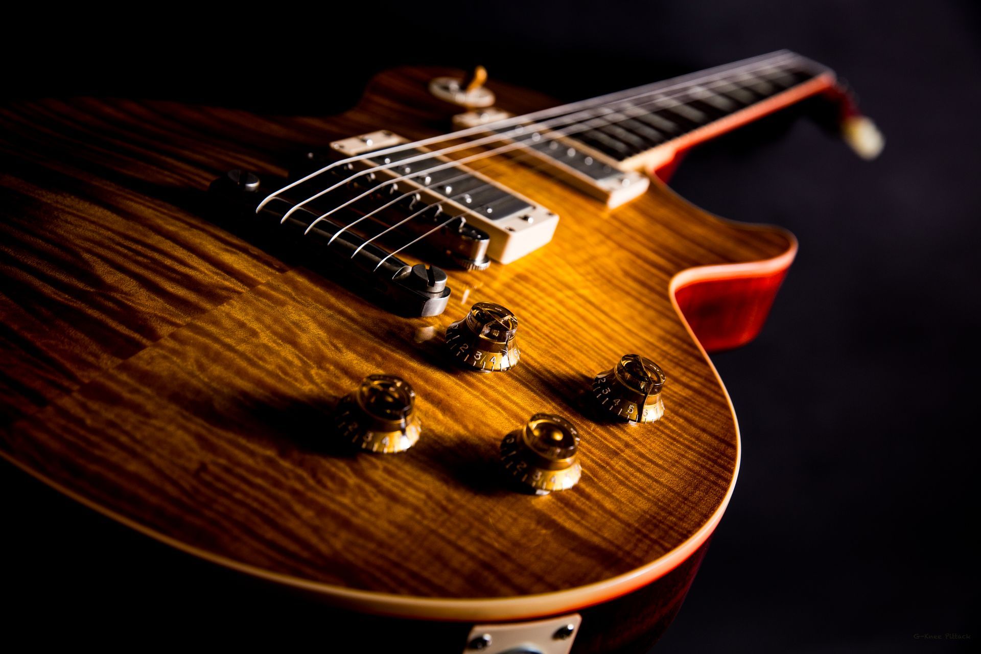 Close-up of a brown, wood-grain electric guitar with knobs and strings, resting on a dark surface.