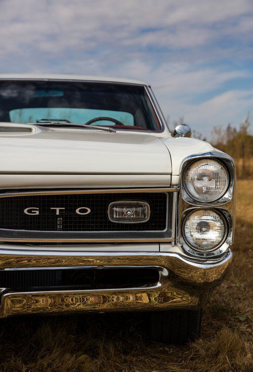 White 1965 Pontiac GTO front view. Classic car with chrome details against a grassy field and blue sky.