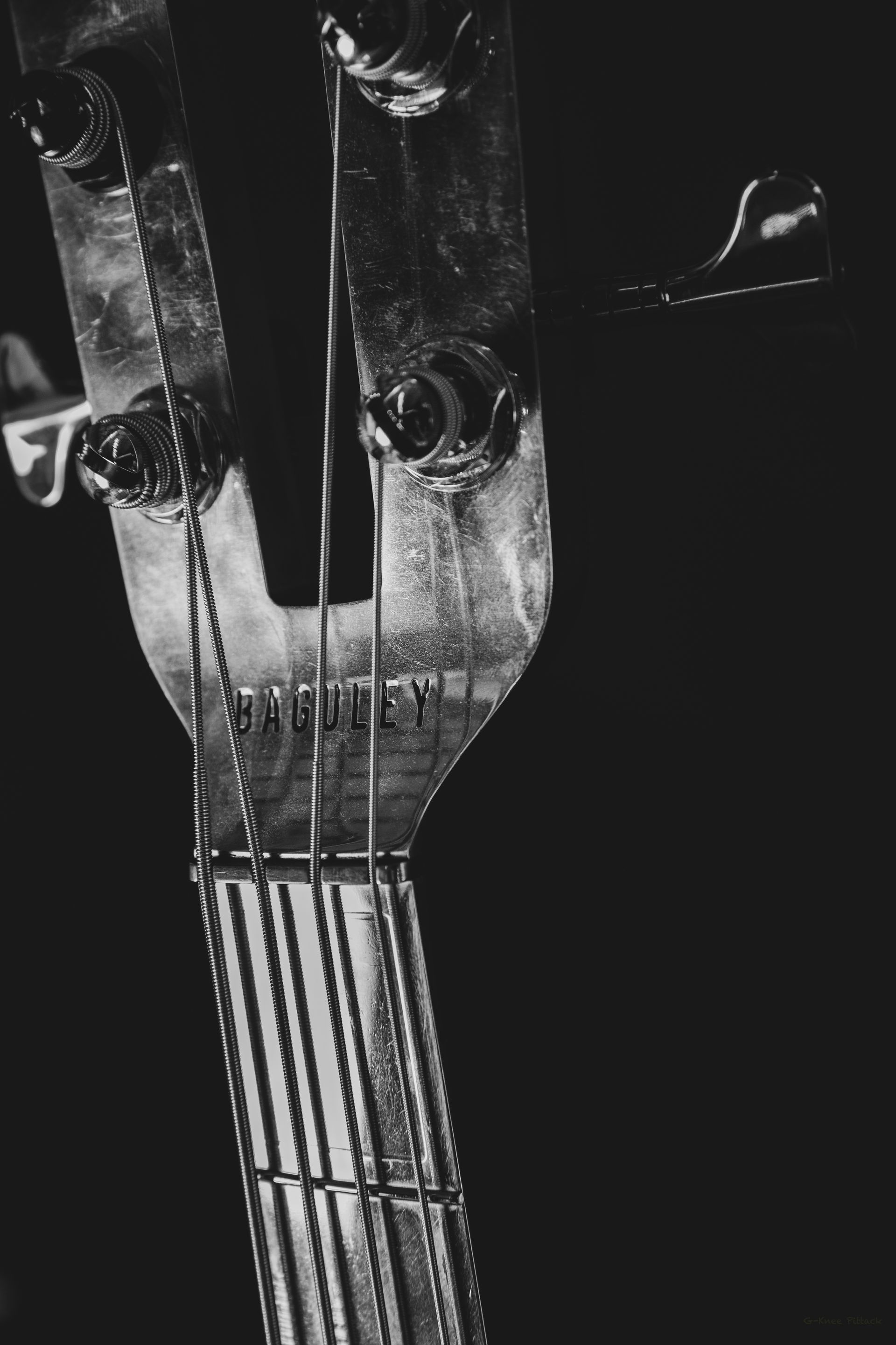Close-up of bass guitar headstock and neck against a dark background. Strings and tuning pegs are visible.