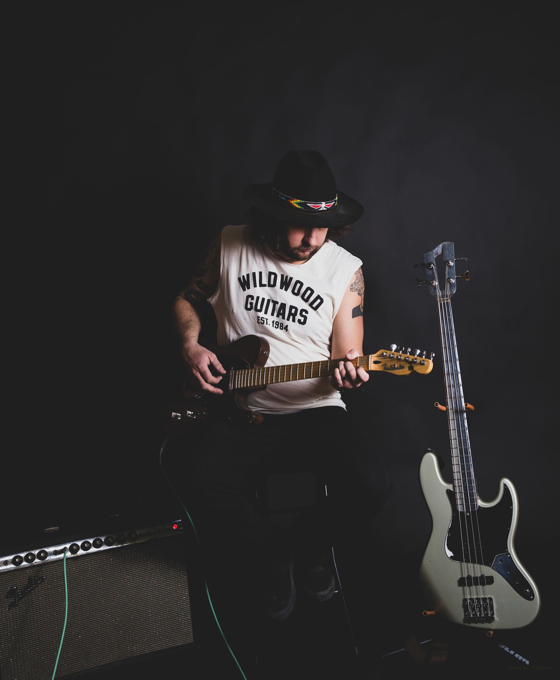 Man playing guitar, wearing a hat and sleeveless shirt. Black background, amp, and bass guitar nearby.