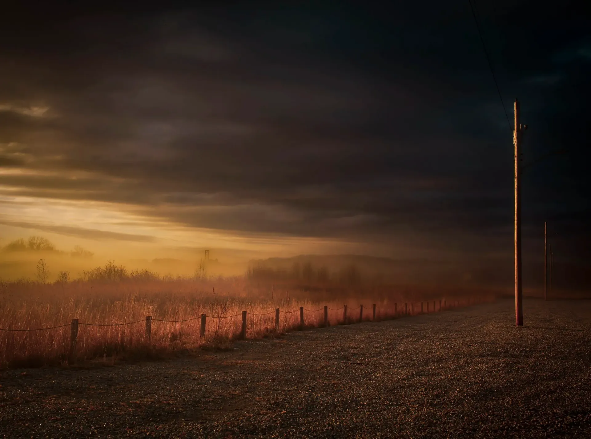 Gloomy rural landscape with a dark sky, misty field, wooden fence, and telephone poles.