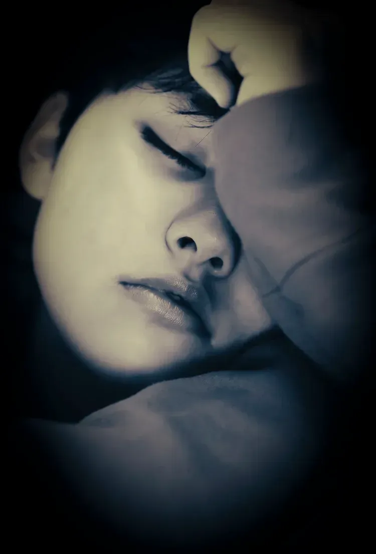 Person with eyes closed, hand on face, possibly sleeping or in thought. Dark lighting.