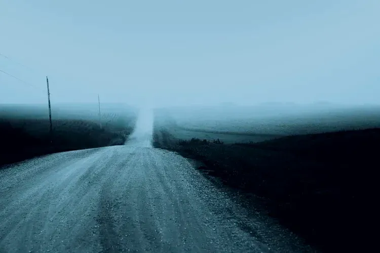 Gravel road disappearing into heavy fog; muted blue tones, rural landscape.