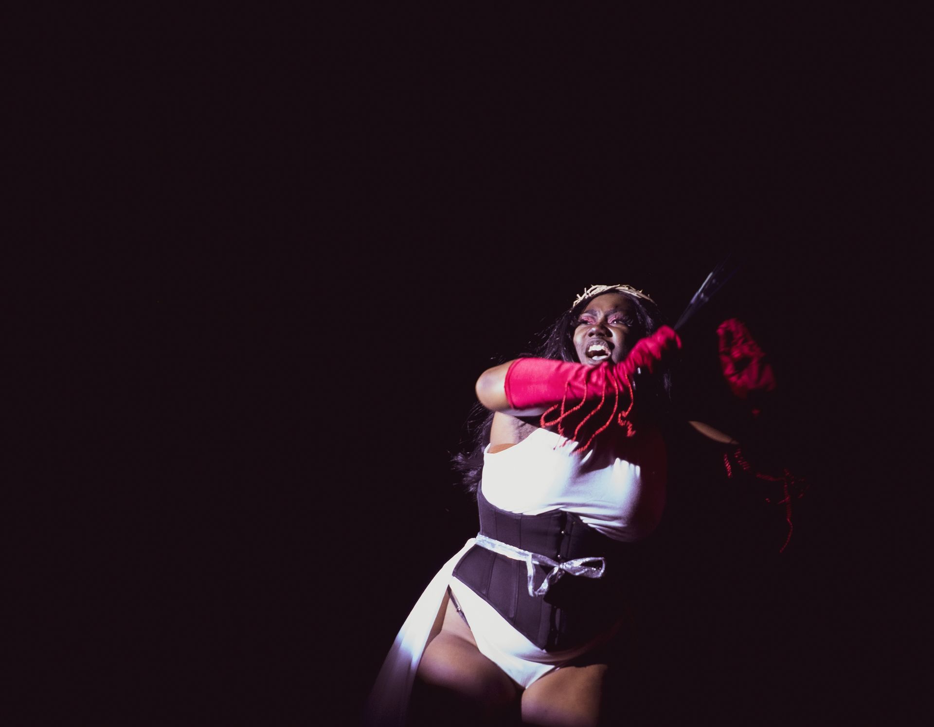 Lizzo performing on stage in a white outfit, red gloves, and a crown. She smiles and gestures with her arms.