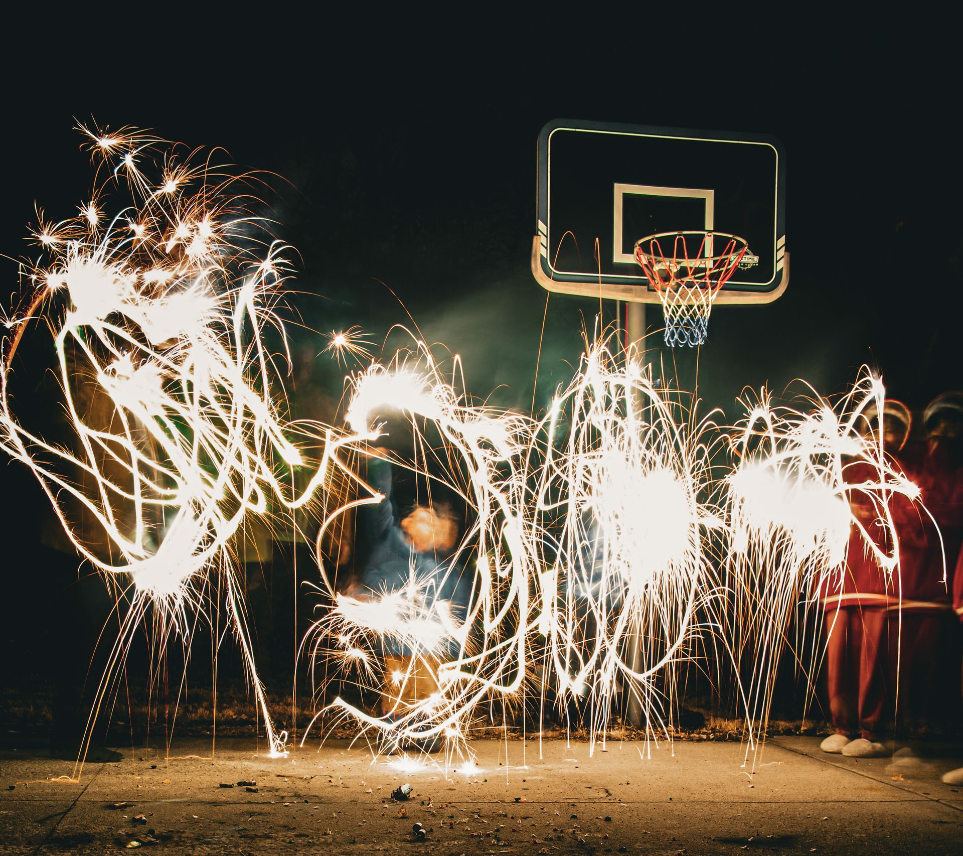 Fireworks light painting around a basketball hoop at night; people in the background.