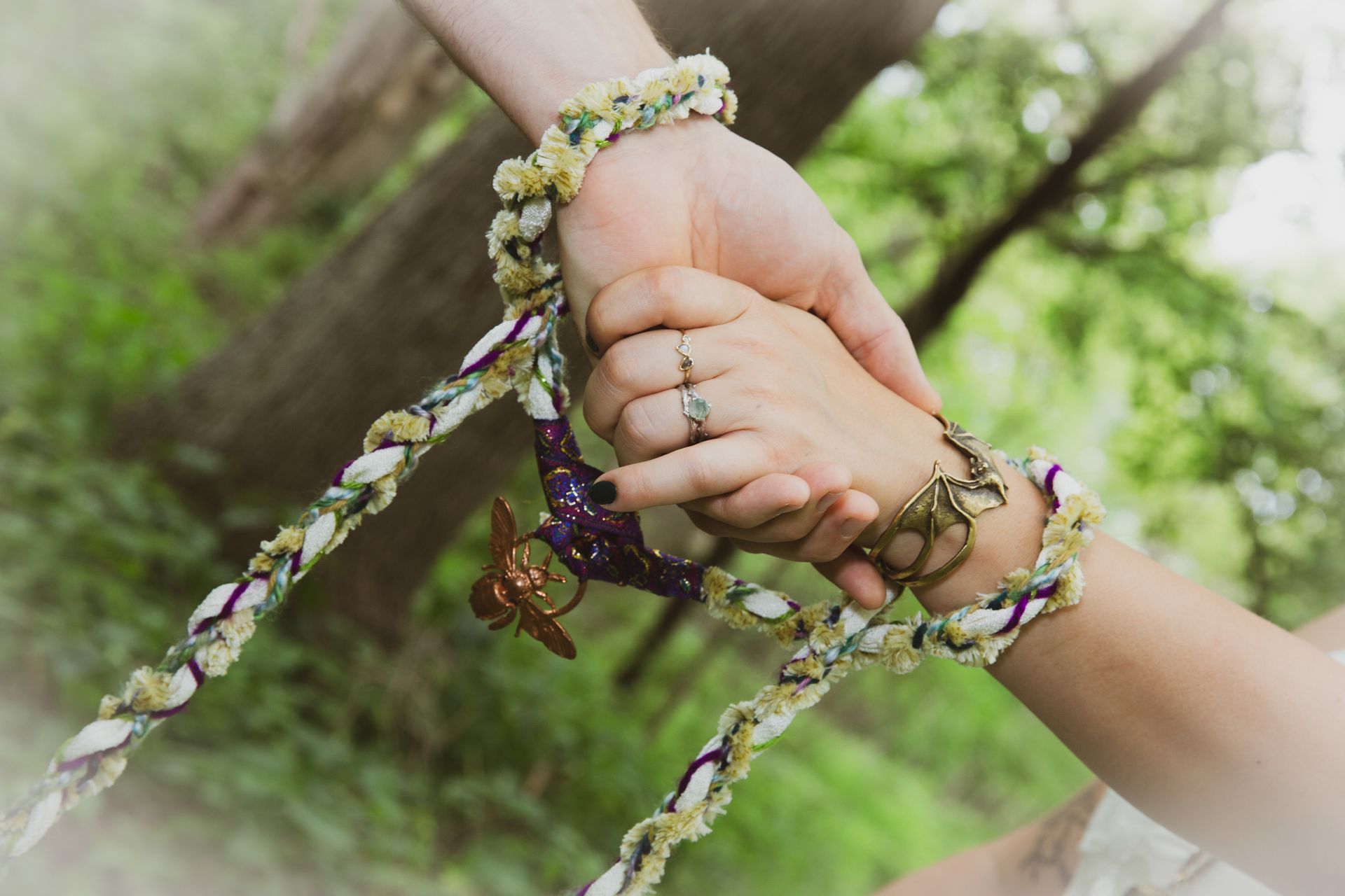 Hands clasped with floral rope in a forest setting; rings are visible.