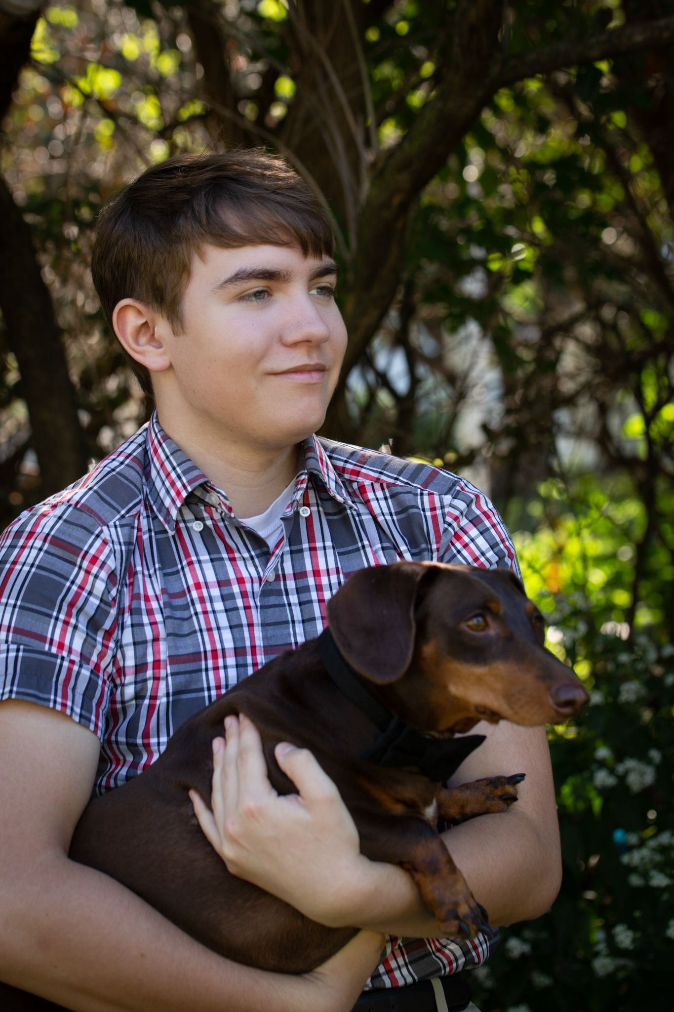 Young man with brown hair, holding a brown dachshund dog, smiles outdoors. He wears a plaid shirt.