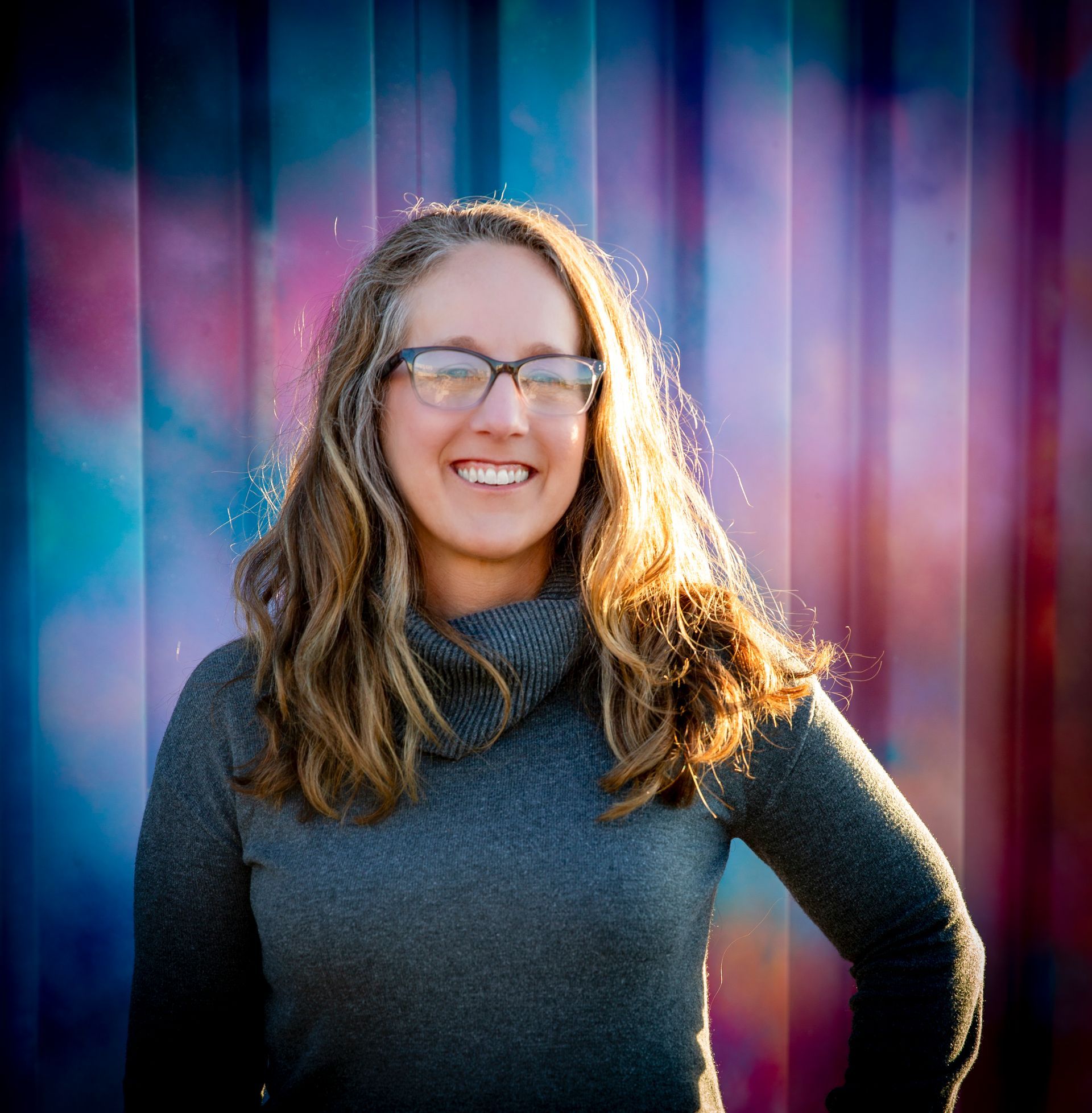 Woman with glasses smiles at the camera in front of a colorful mural; she wears a gray sweater.