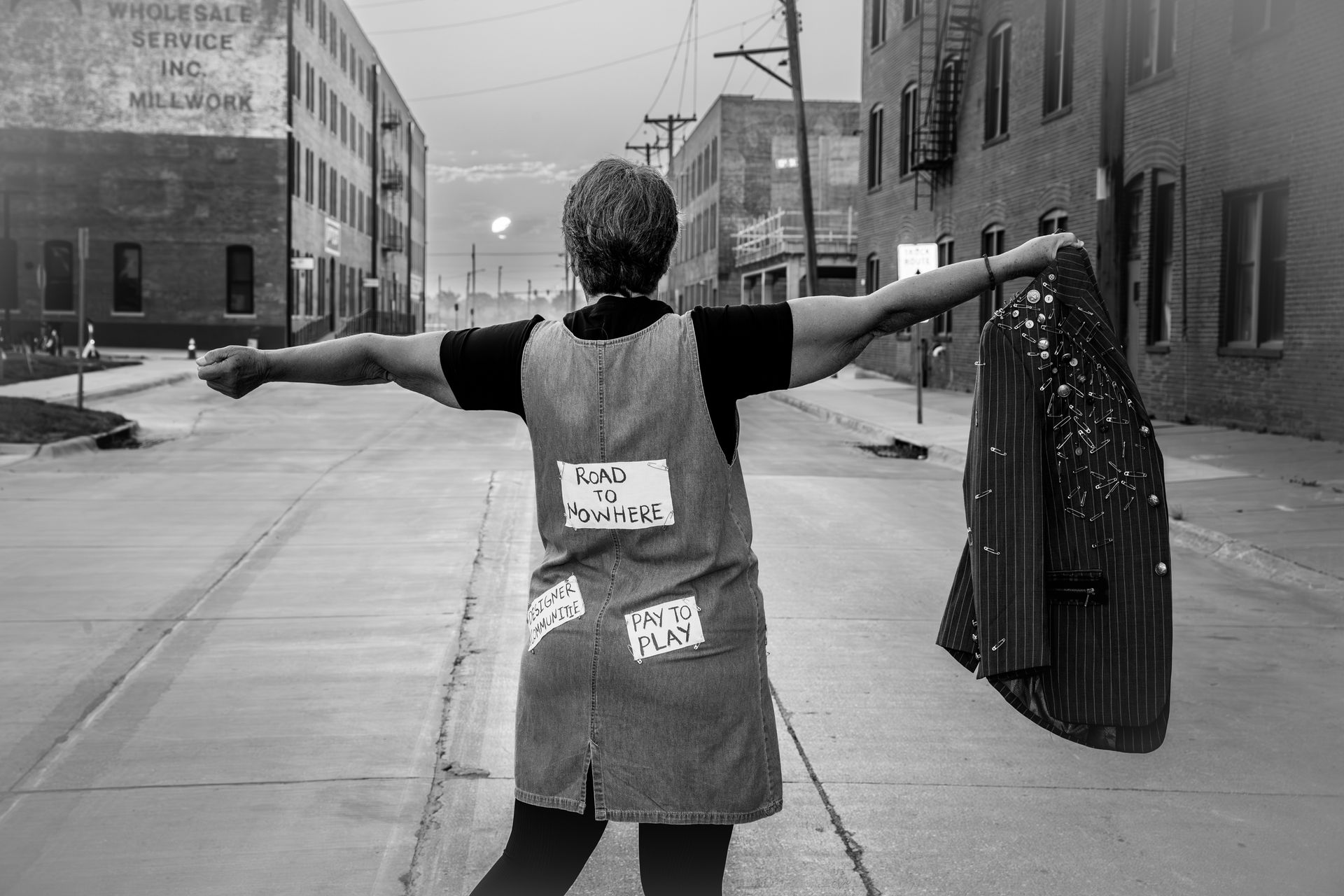 Woman in overalls with patches stands in a city street, arms outstretched, holding a sparkling jacket.