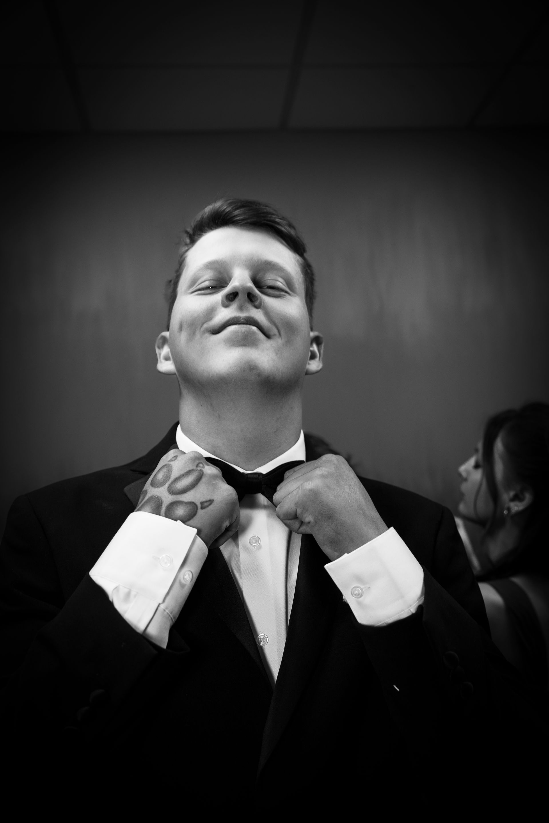 Man in tuxedo adjusting bow tie, smiling. Black and white photo.