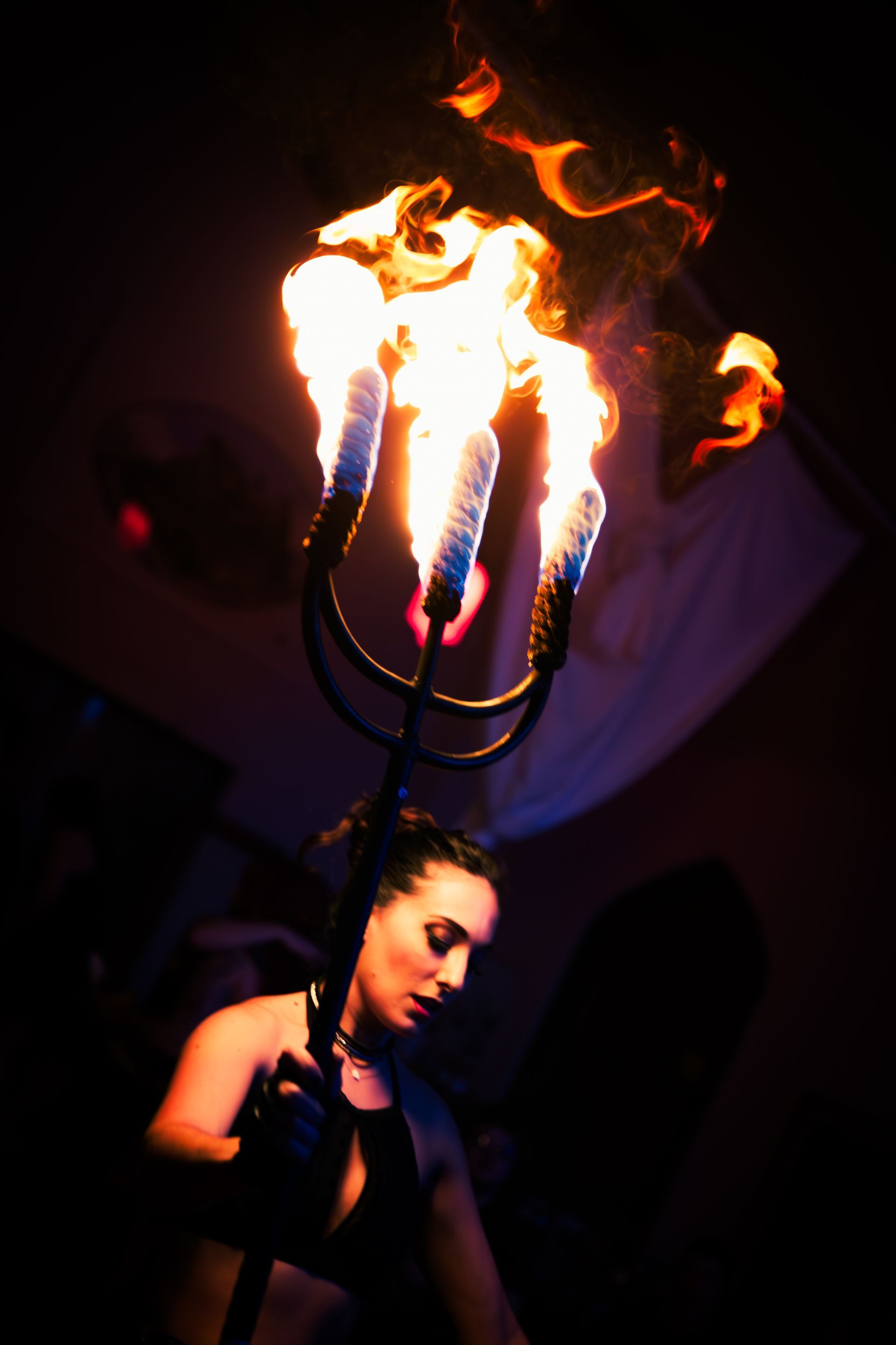 Woman holding a flaming trident aloft, face lit by fire, in a dimly lit space.