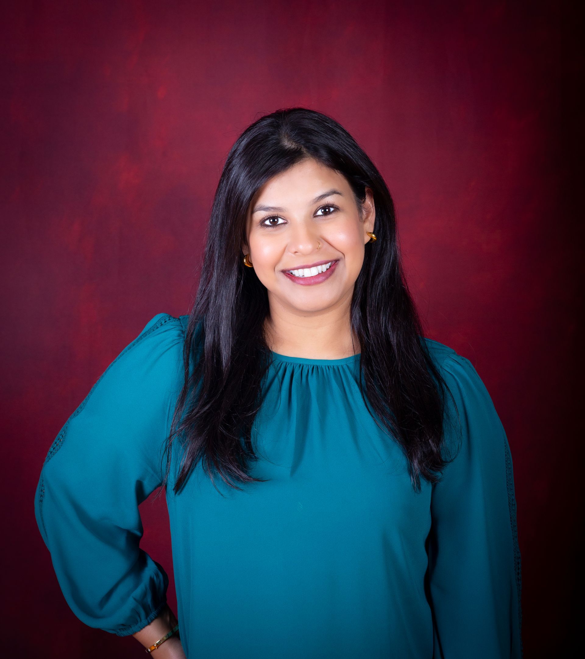 Woman with dark hair smiles, wearing a teal blouse, against a red backdrop.