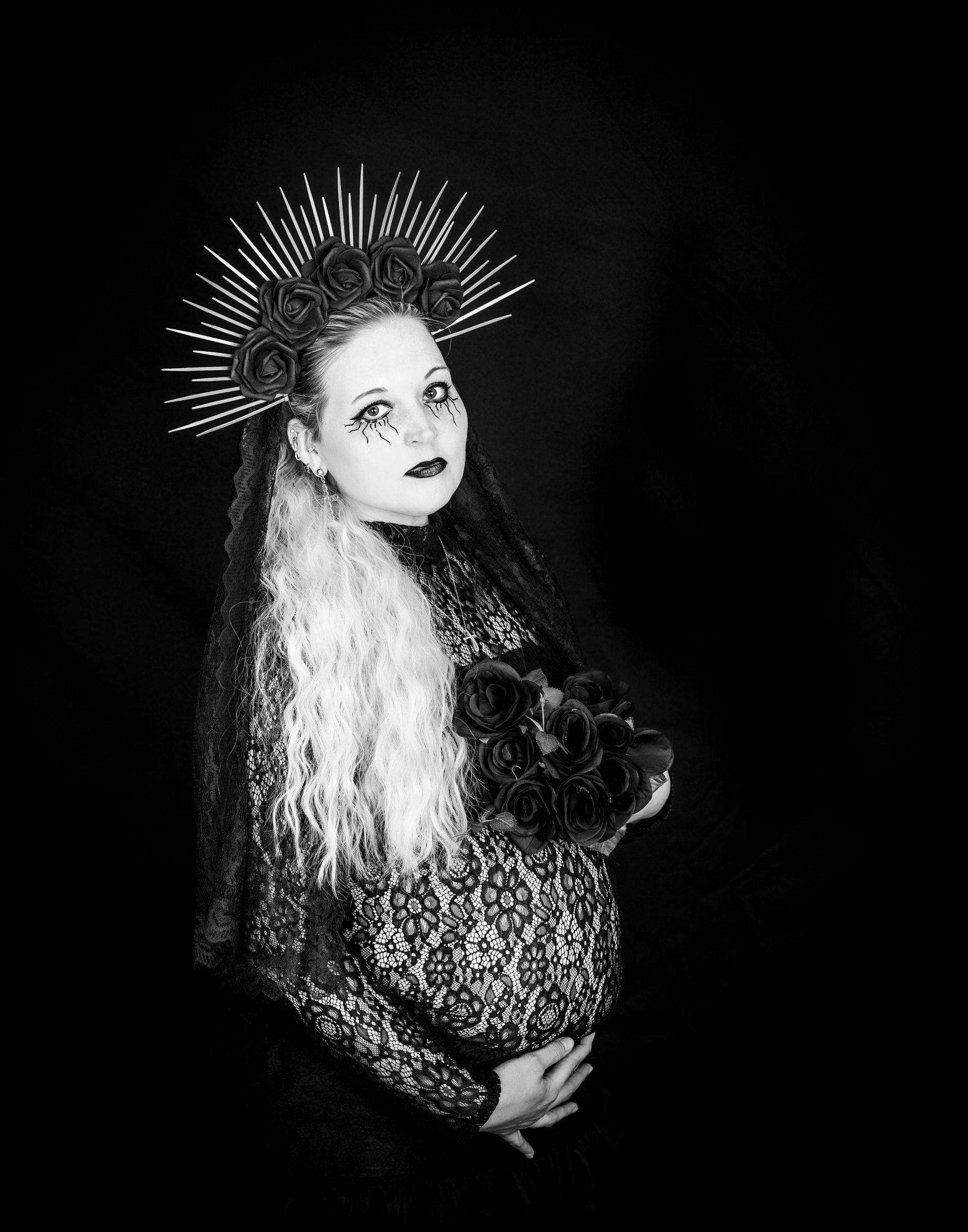 Pregnant woman in goth attire, holding black roses, wearing ornate headpiece, black and white.