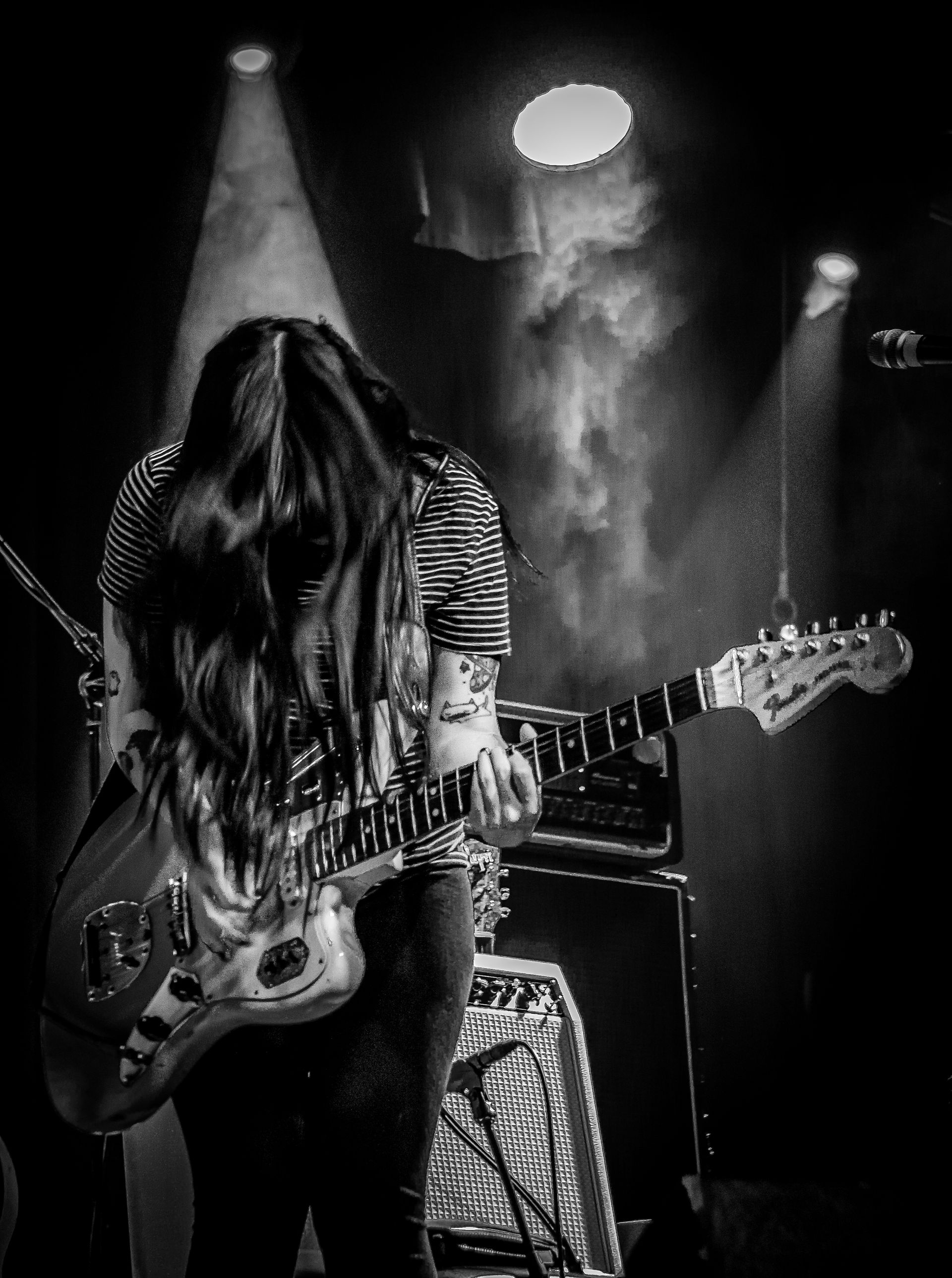 Woman playing electric guitar onstage, black and white. Stage lights, smoky atmosphere.