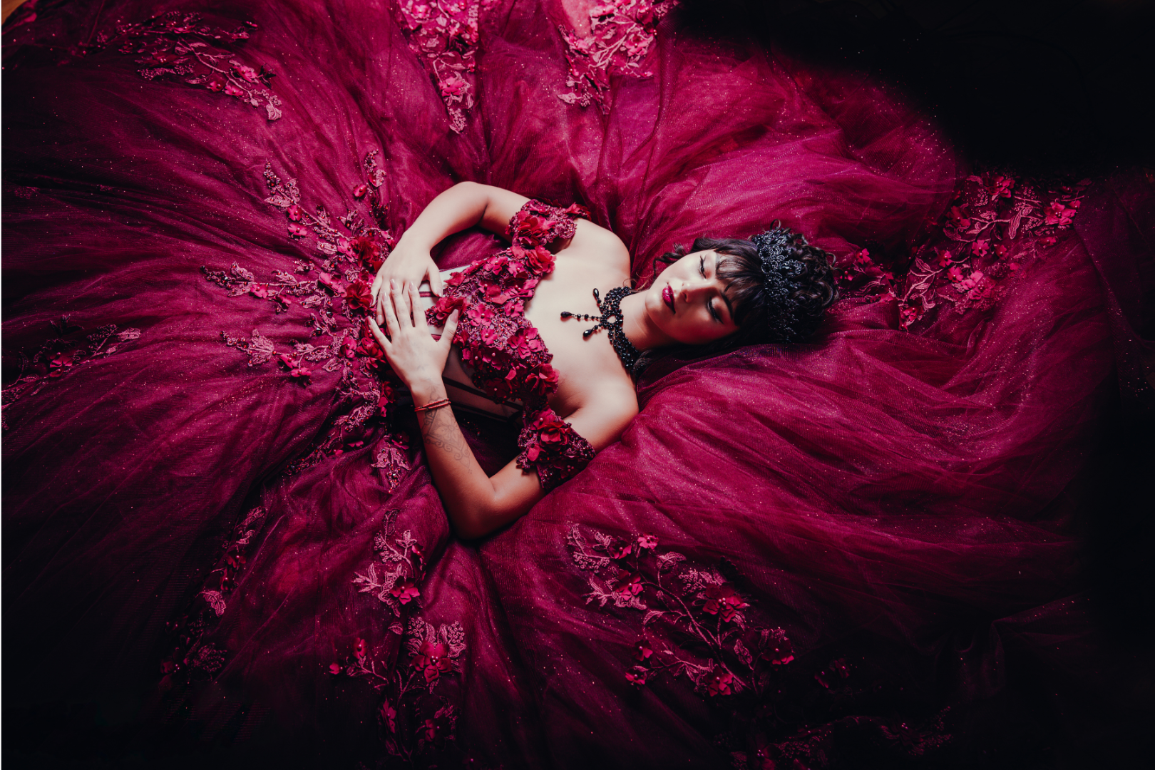 Woman in a glittering, maroon ball gown lies on a dark surface, arms crossed.