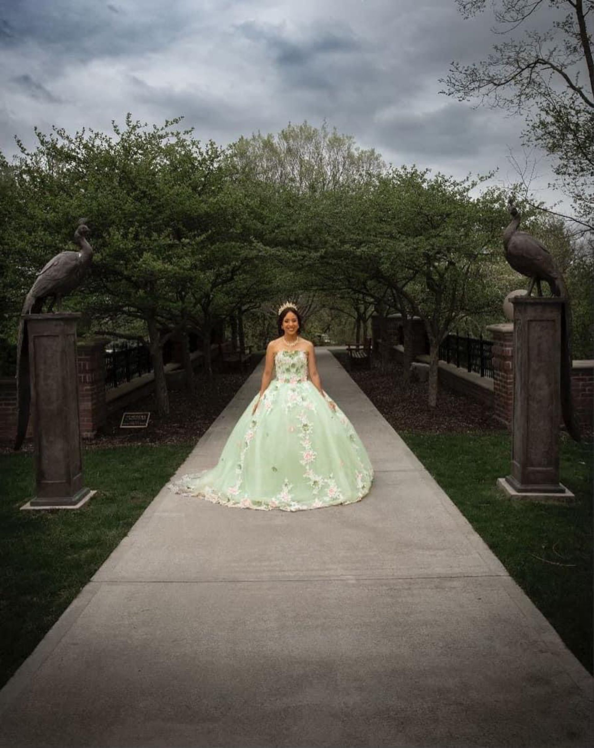 Woman in a light green quinceañera dress poses on a pathway flanked by sculptures and trees, under an overcast sky.