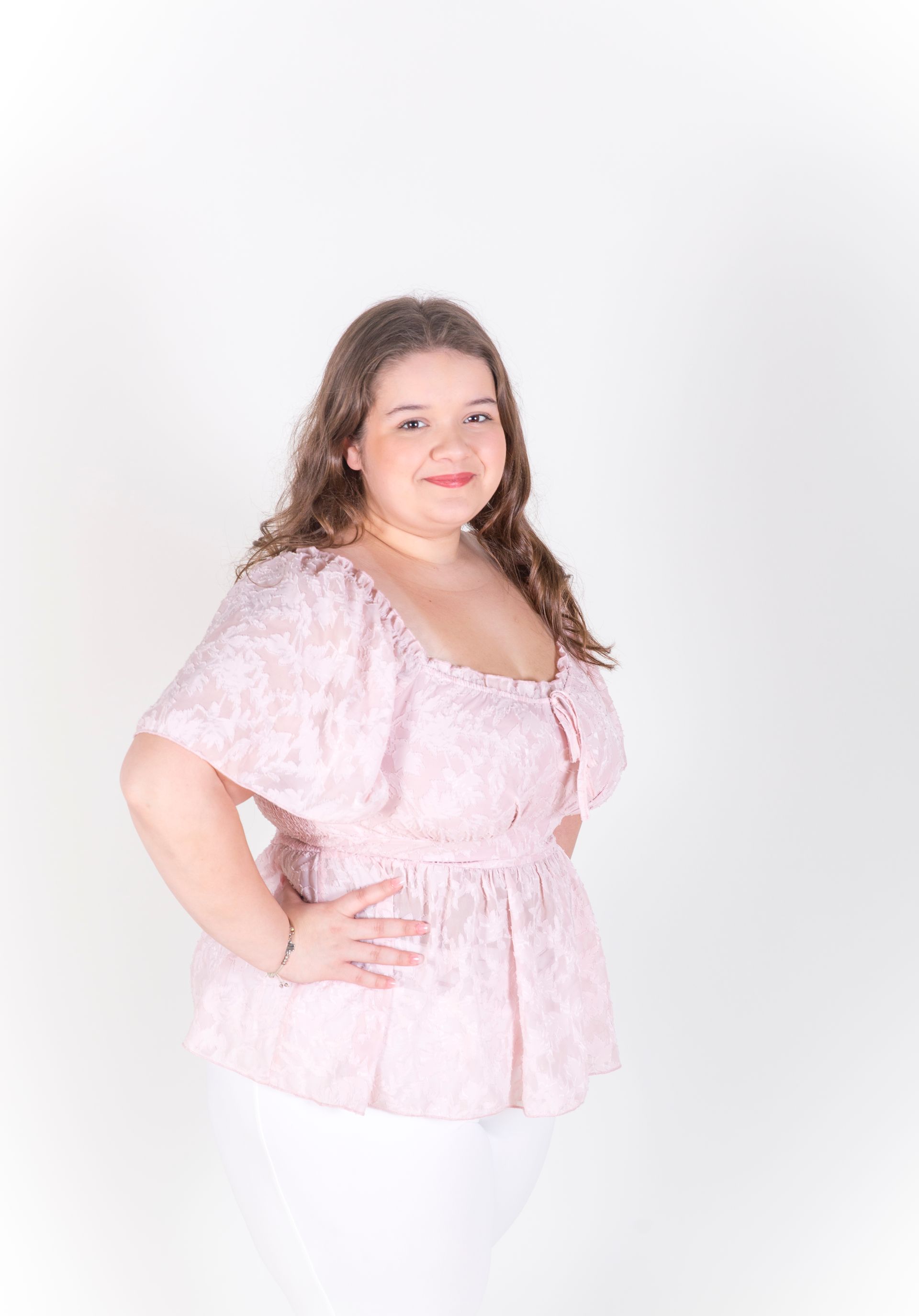 Woman with curly hair in a pink top and white pants, smiling, hands on hips, against a white backdrop.