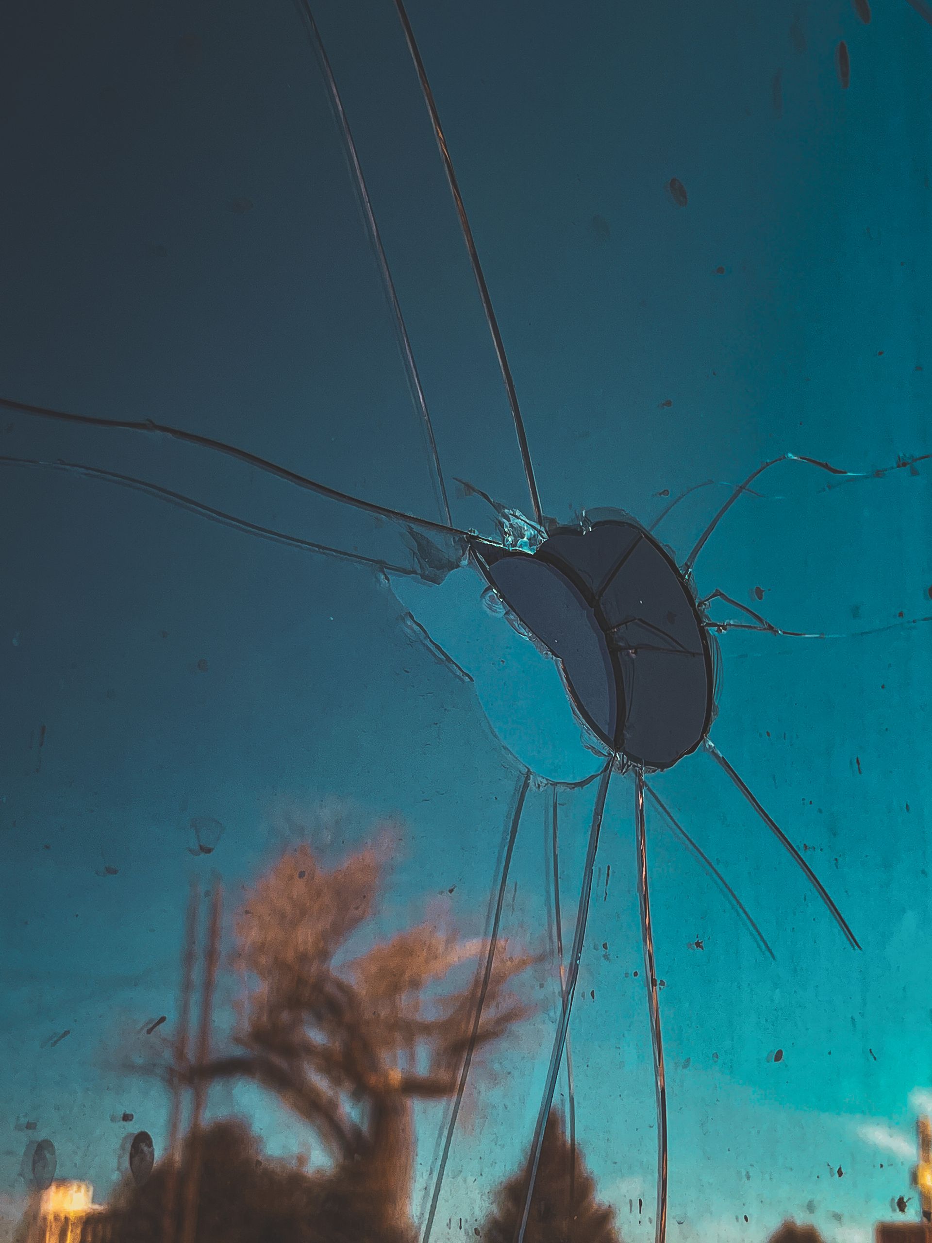 Cracked glass with a dark hole in the center, tree and blue sky visible through the cracks.