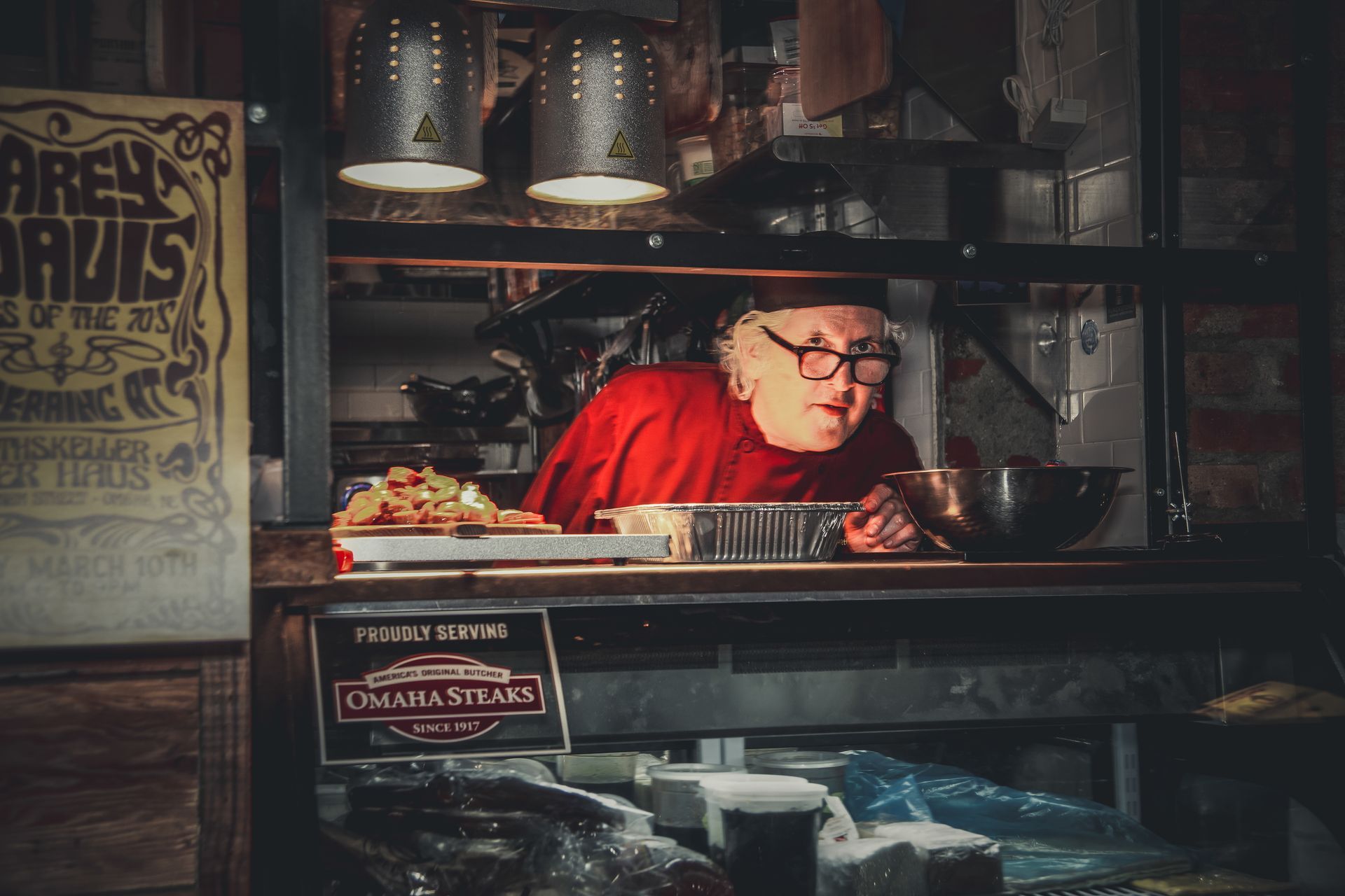 Chef in red shirt leans over a counter in a dimly lit restaurant kitchen. A poster and food are visible.