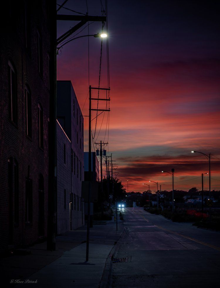 Street scene at dusk; red and purple sky. Buildings, power lines, streetlights illuminate the road.