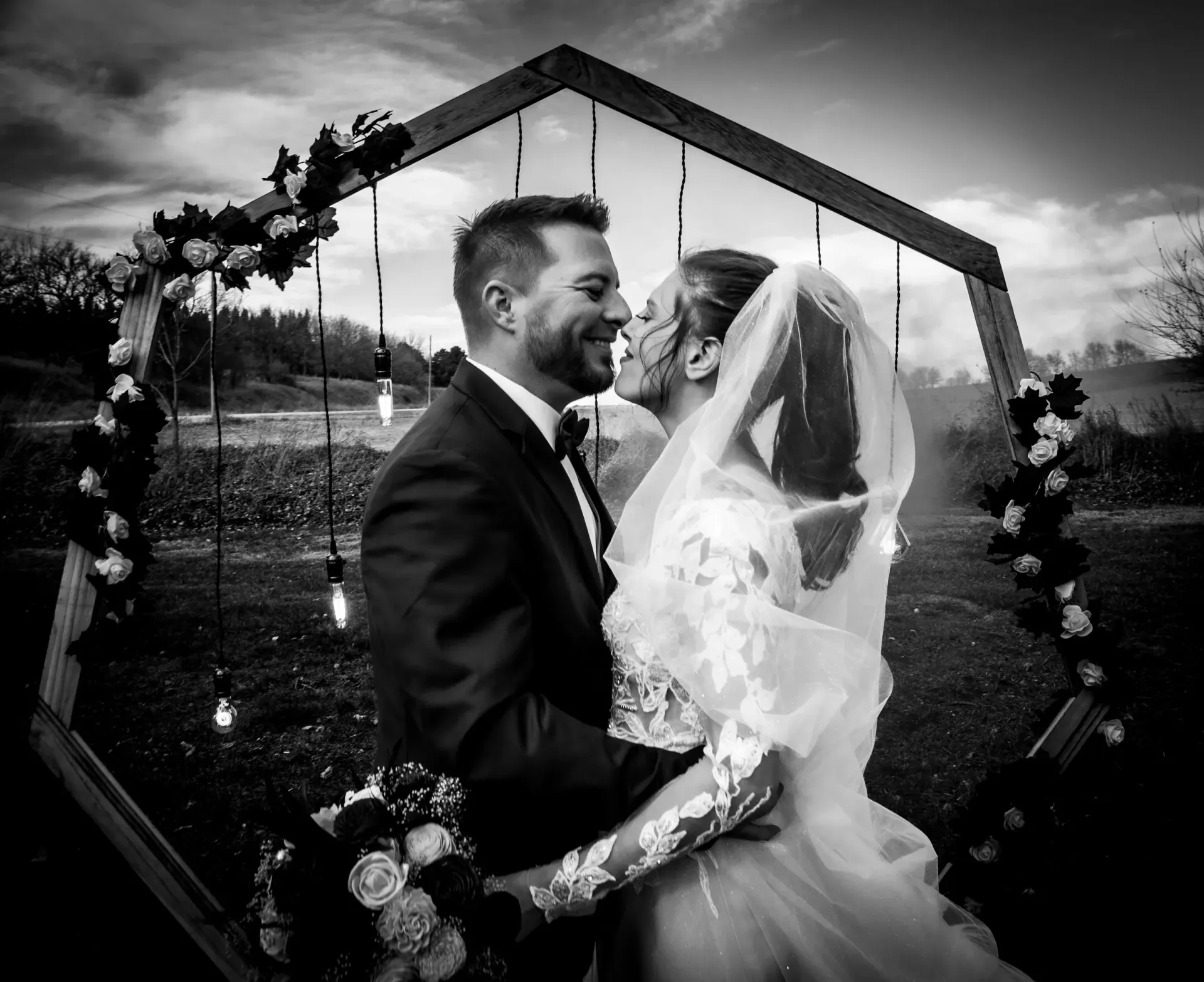 Bride and groom embrace, kissing, in front of decorated wooden arch at outdoor wedding. Black and white.