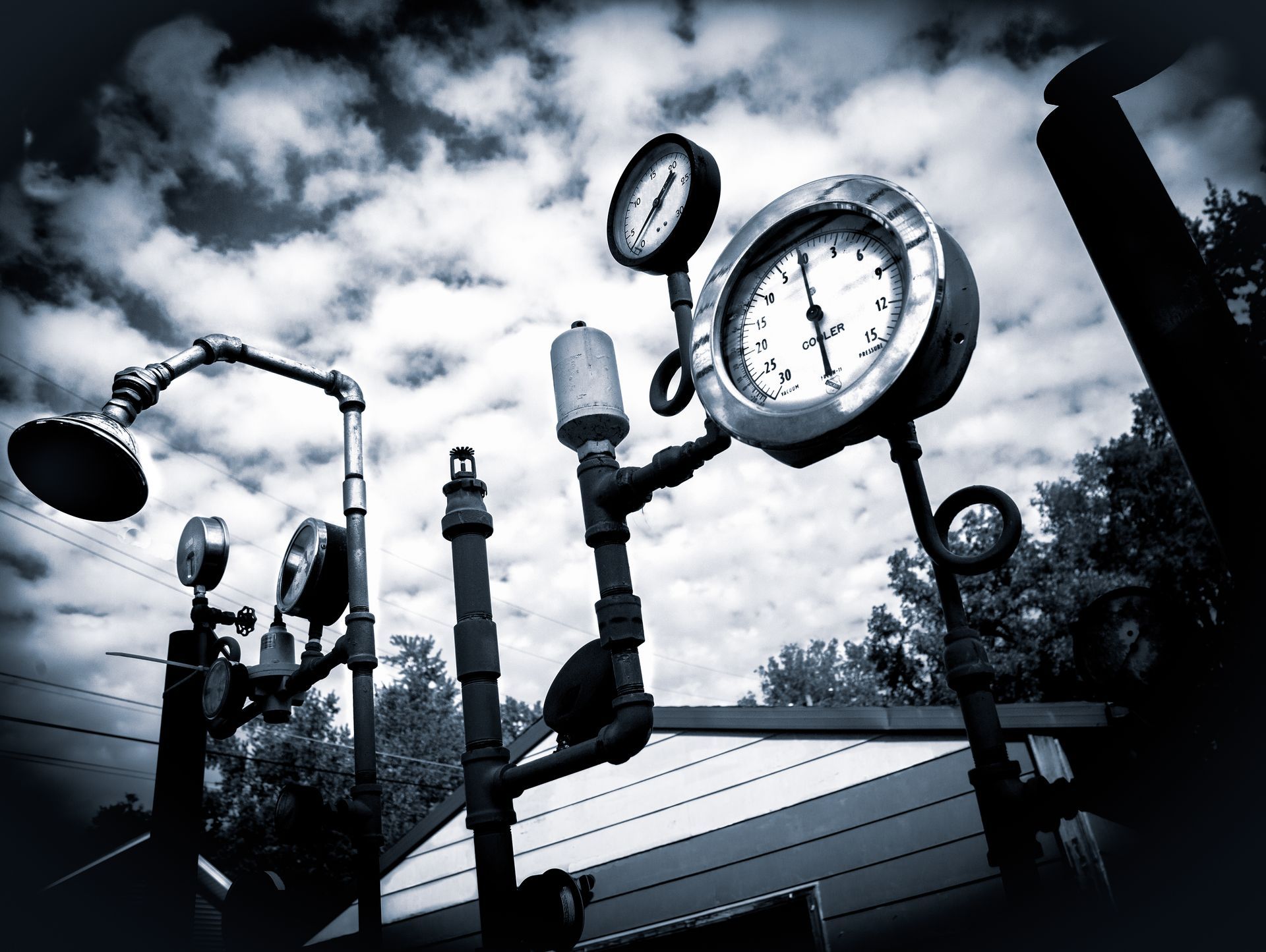 Black and white image of pipes with pressure gauges against a cloudy sky.