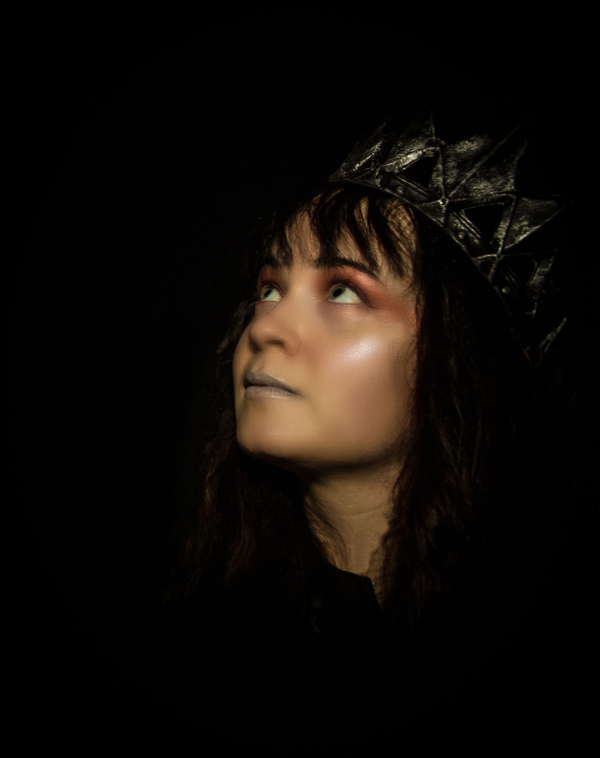 Woman with a silver crown looking upwards, face lit against a dark background.