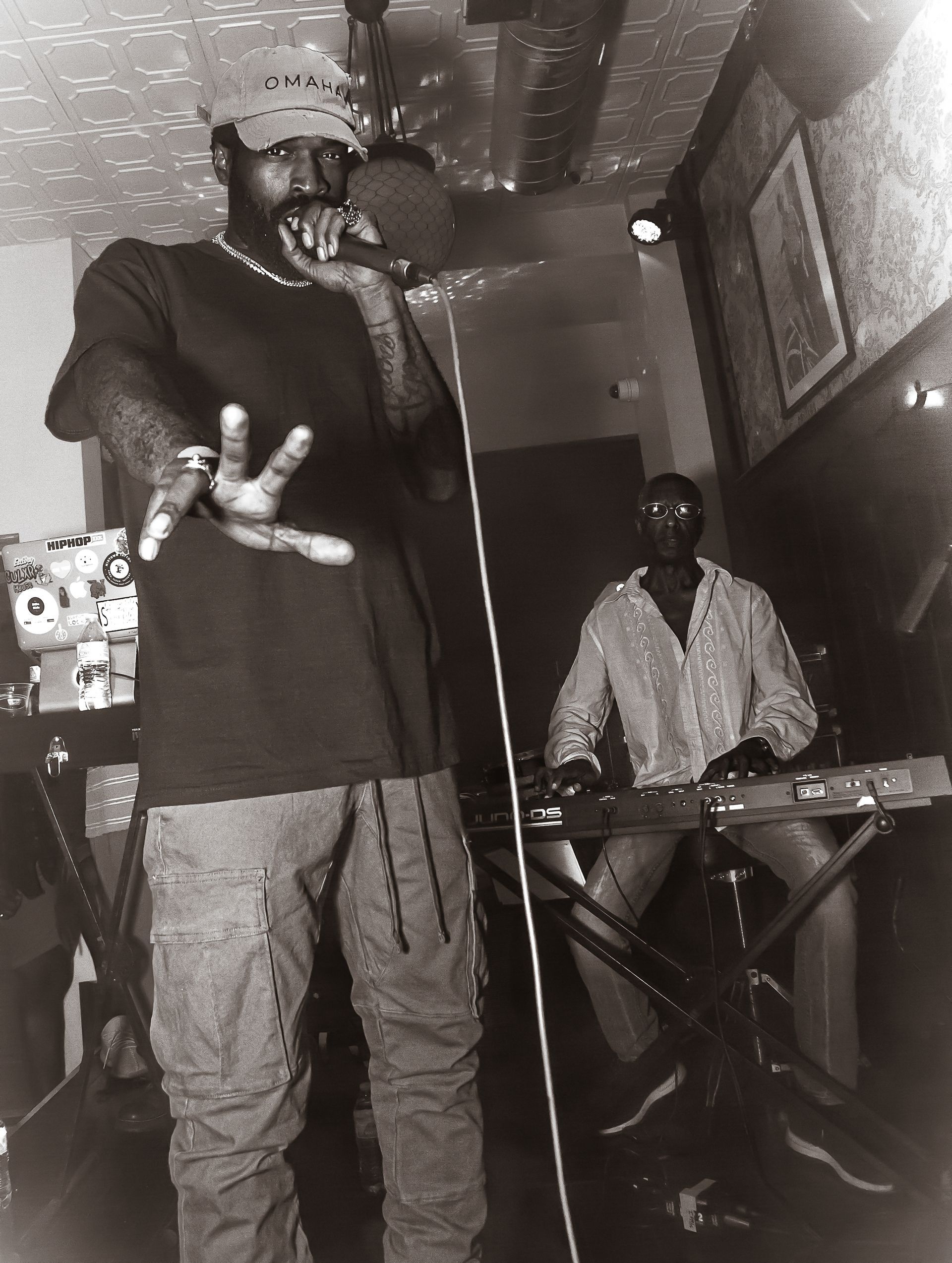 Black and white photo of a Black man rapping into a mic. Another man plays a keyboard in the background.