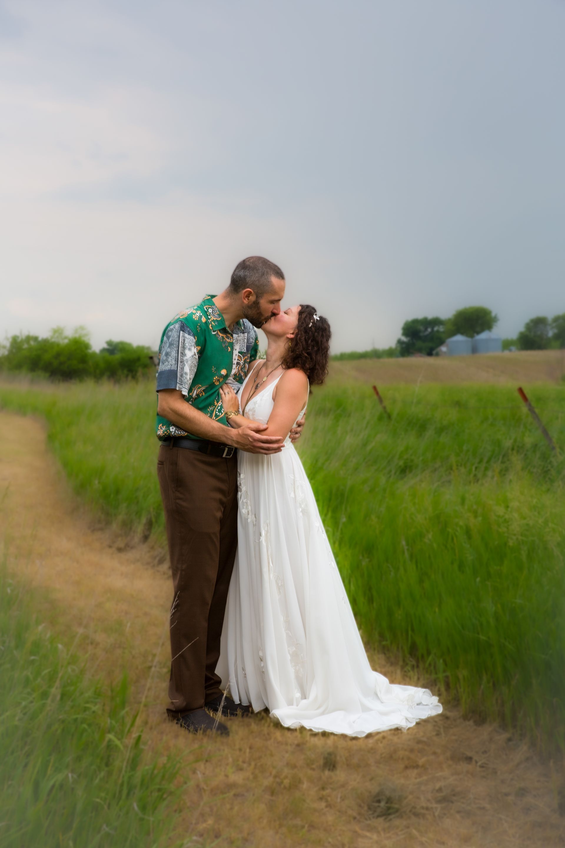 Couple kissing, bride in white dress, groom in green shirt, standing in a grassy field.