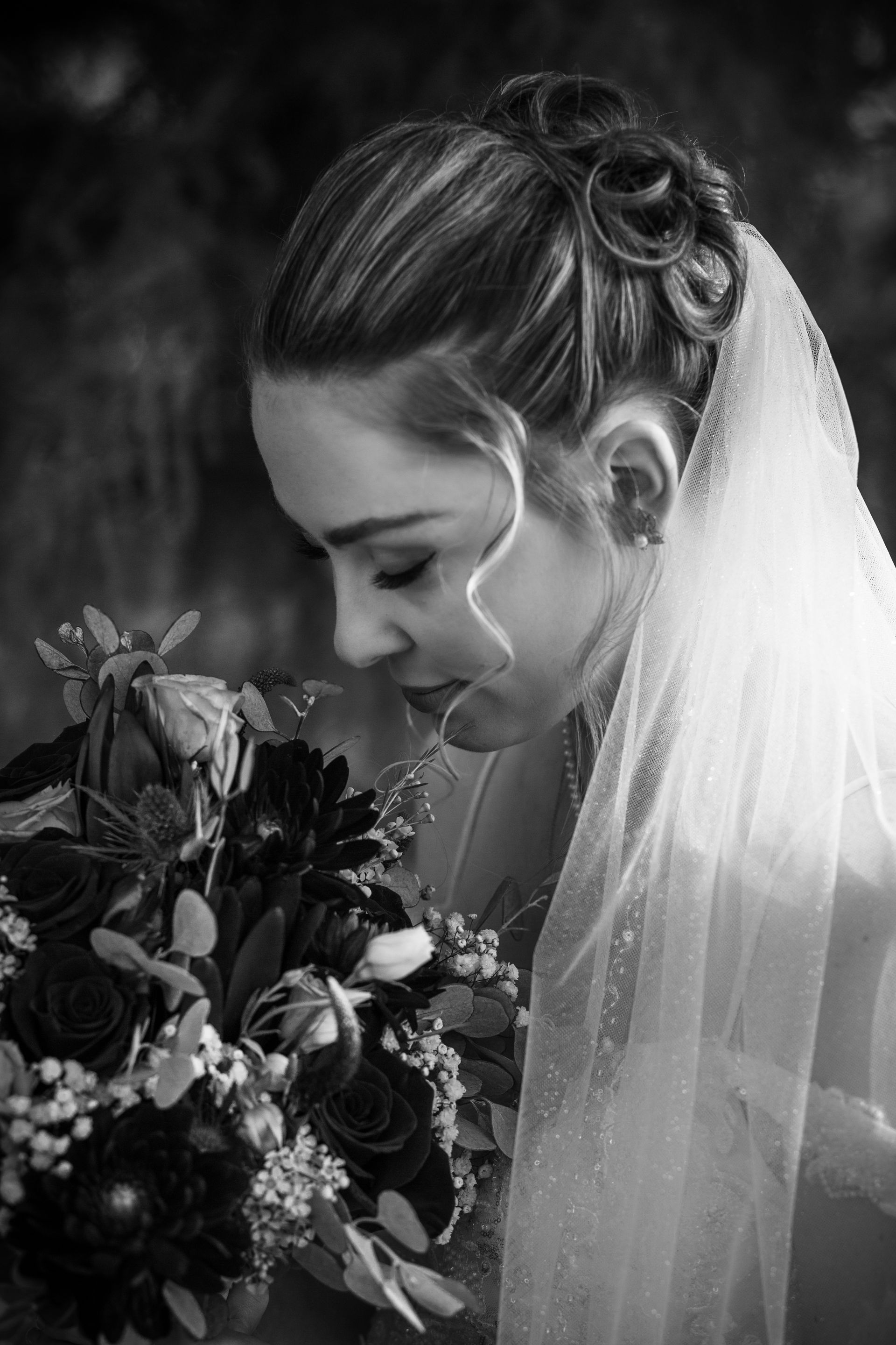 Bride in profile, looking down at a dark floral bouquet, with veil and updo in black and white.