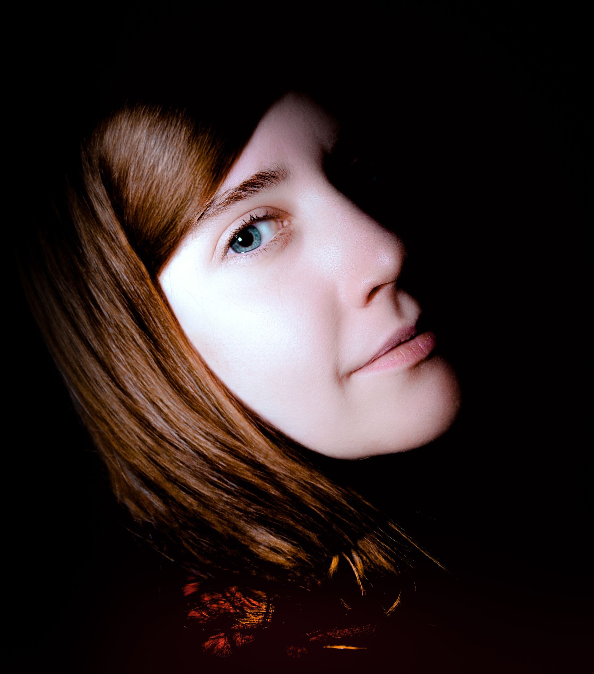 Woman's face in profile, fair skin and light blue eyes, framed by brown hair against a black background.
