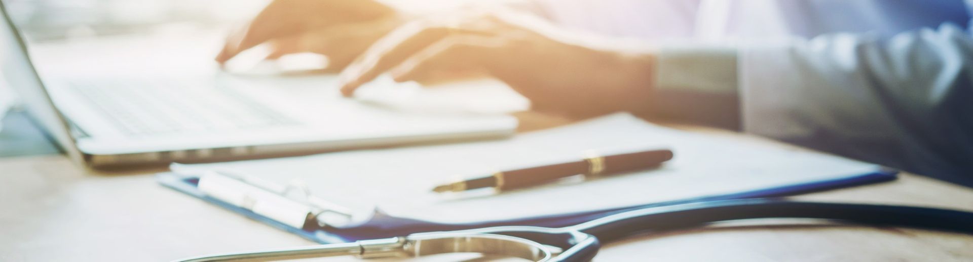 A person types on a laptop next to a clipboard with a pen and a stethoscope on a desk, representing a medical setting.