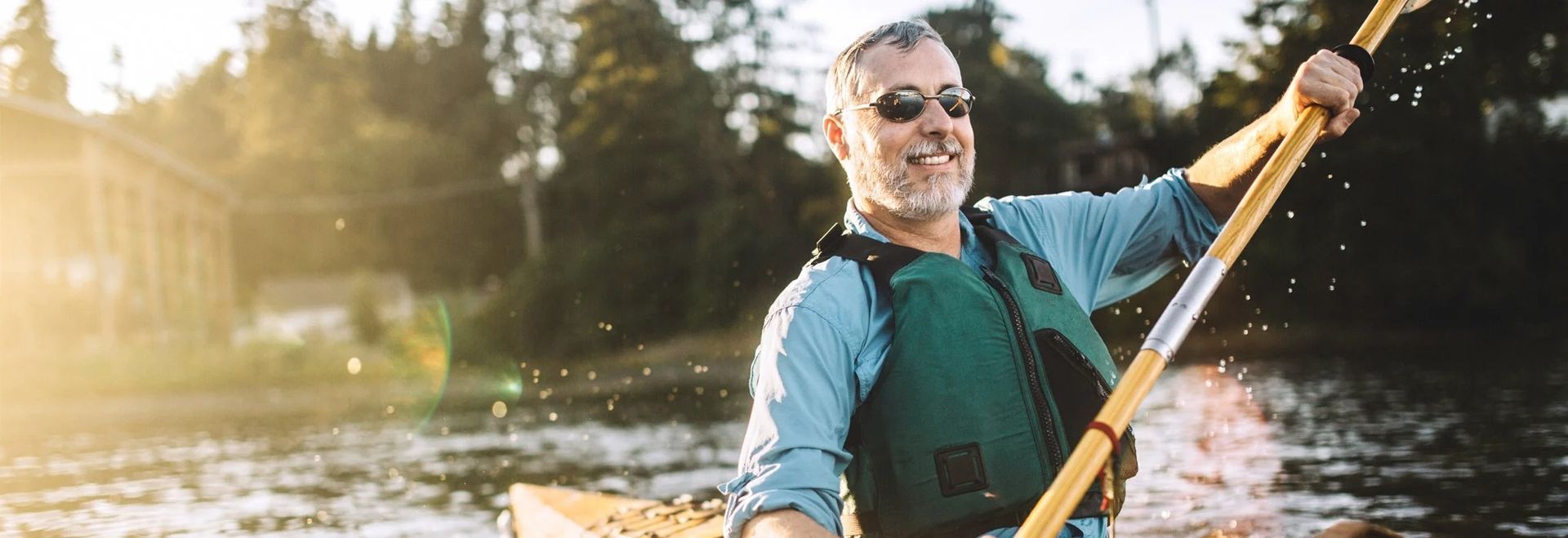 A smiling person in a life vest kayaking on a river during a sunny, golden-hour afternoon.