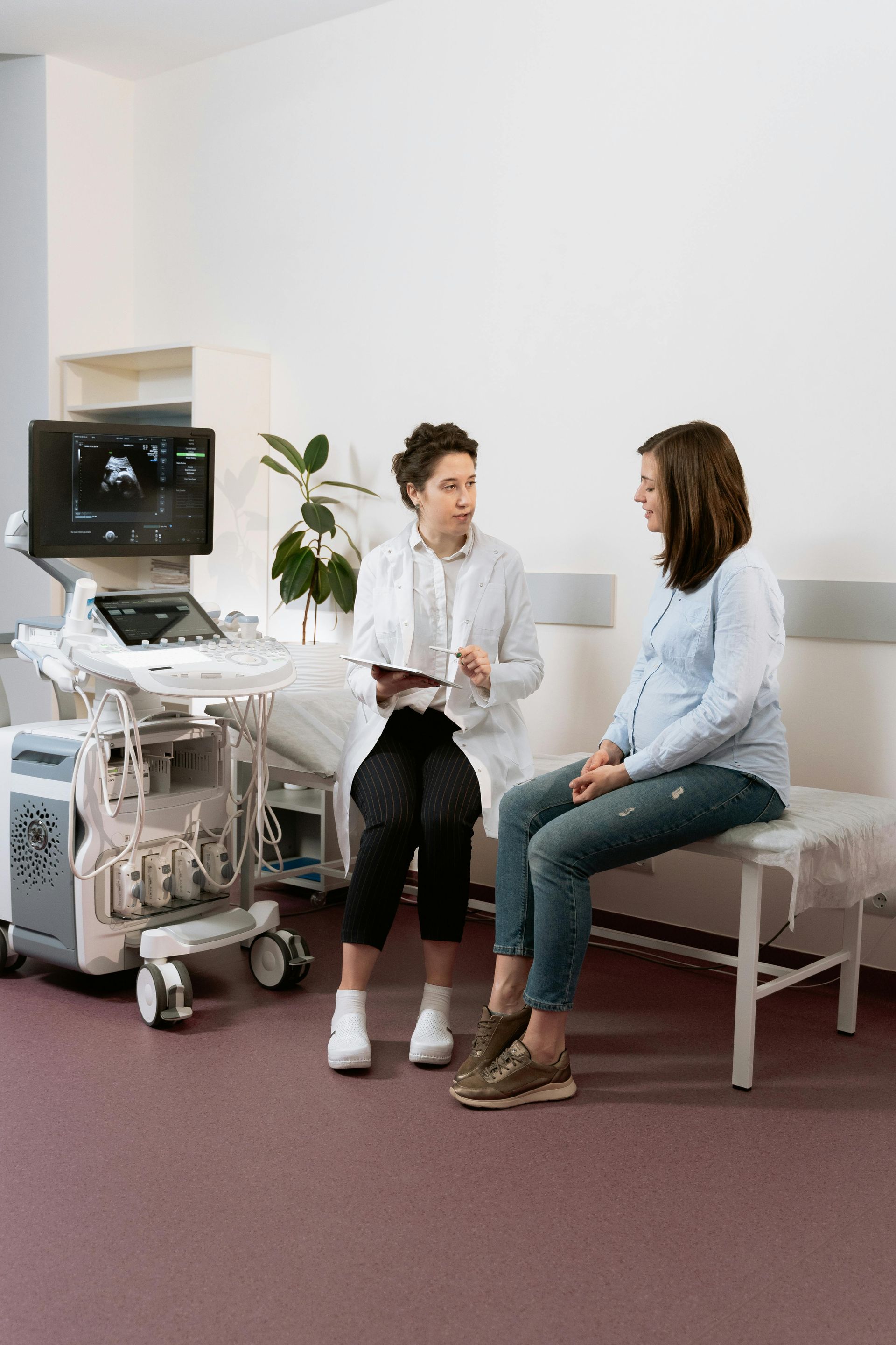 A medical professional in a lab coat talks to a pregnant patient in a clinic next to an ultrasound machine.
