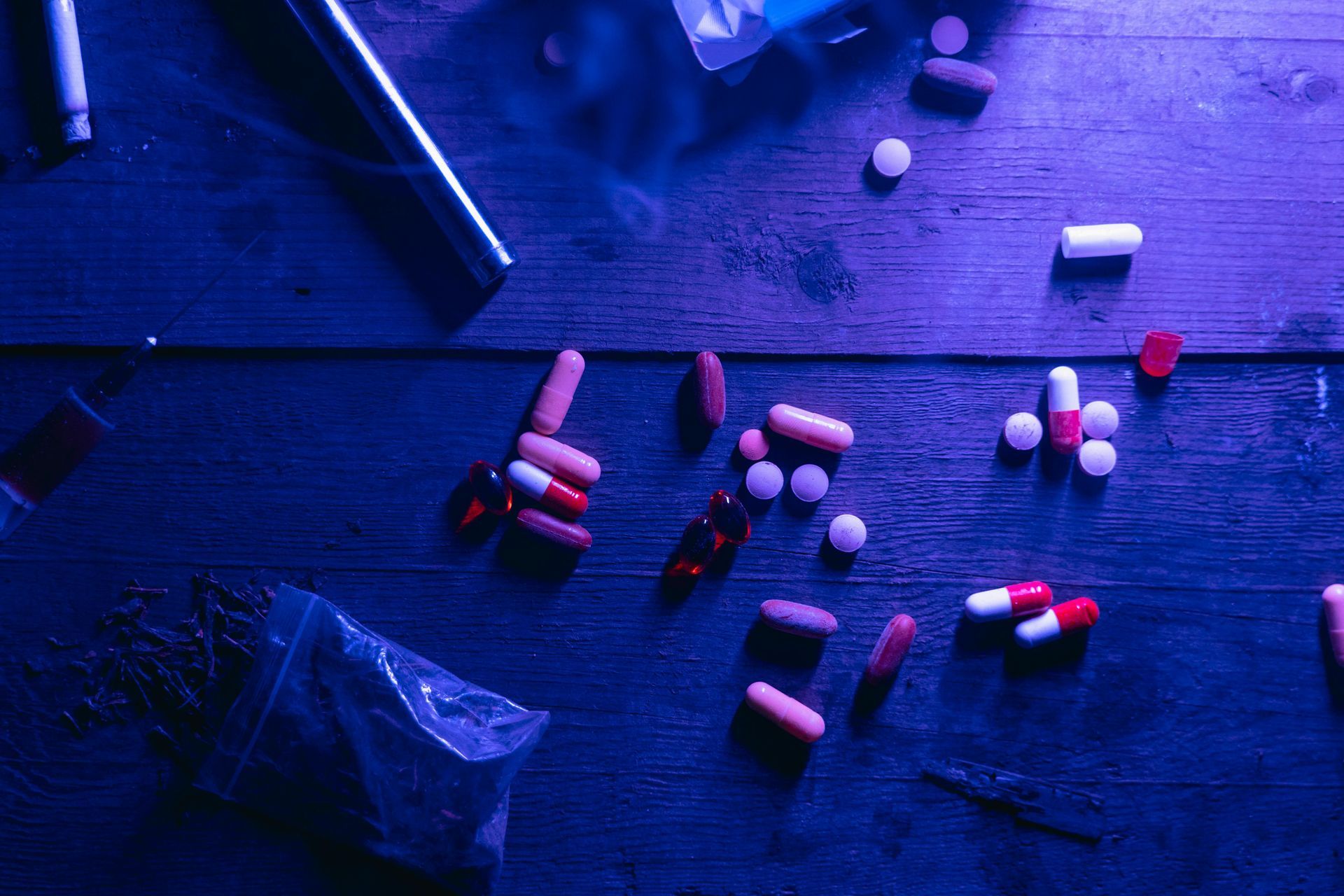 A variety of scattered pills, capsules, and a small plastic bag on a dark wooden surface illuminated in blue light.