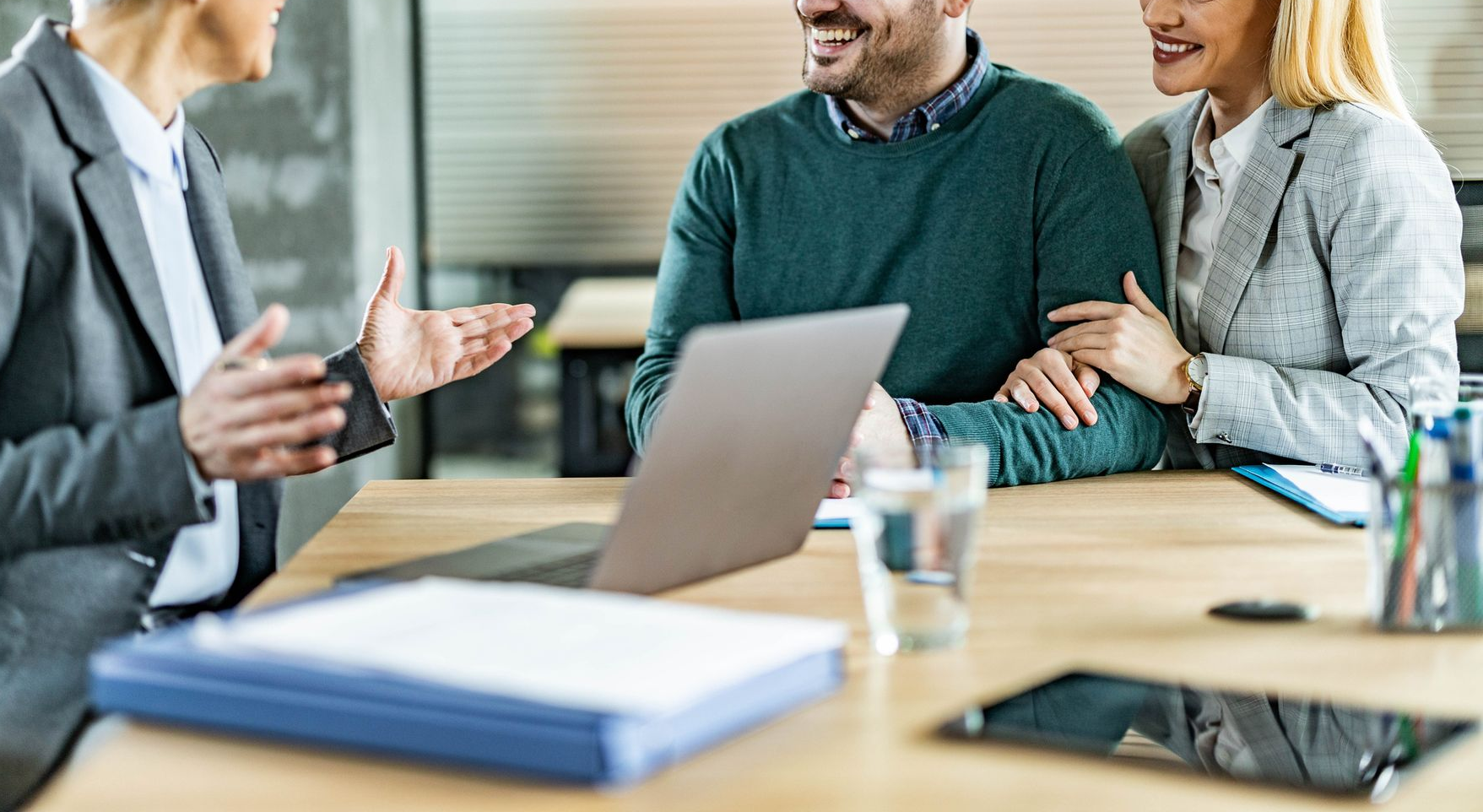 Una donna in giacca e cravatta dà consigli a una coppia sorridente seduta a una scrivania con un computer portatile, documenti e un tablet.