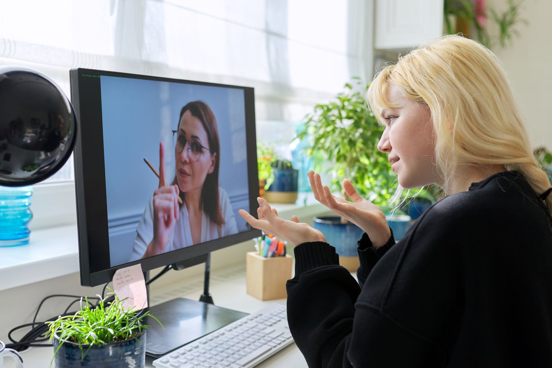 Woman on video call gestures, looking at a monitor displaying another woman pointing a finger, in a home office setting.