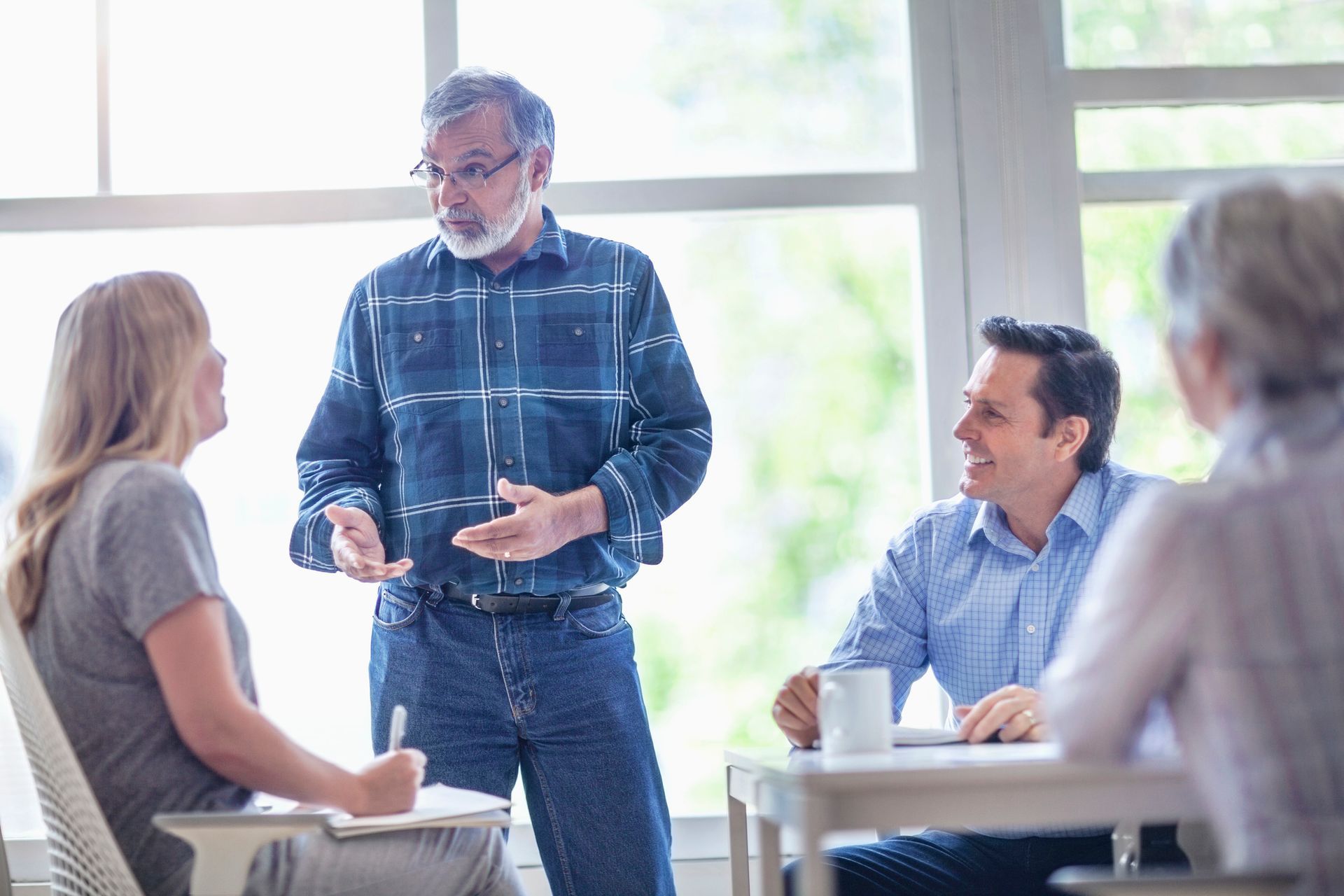 Group of people in a meeting, one man standing and speaking, others seated at a table.