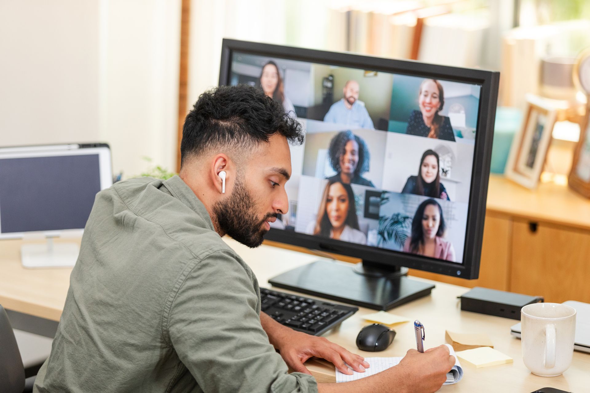 Man in a video conference, writing notes at a desk with a computer displaying other participants.