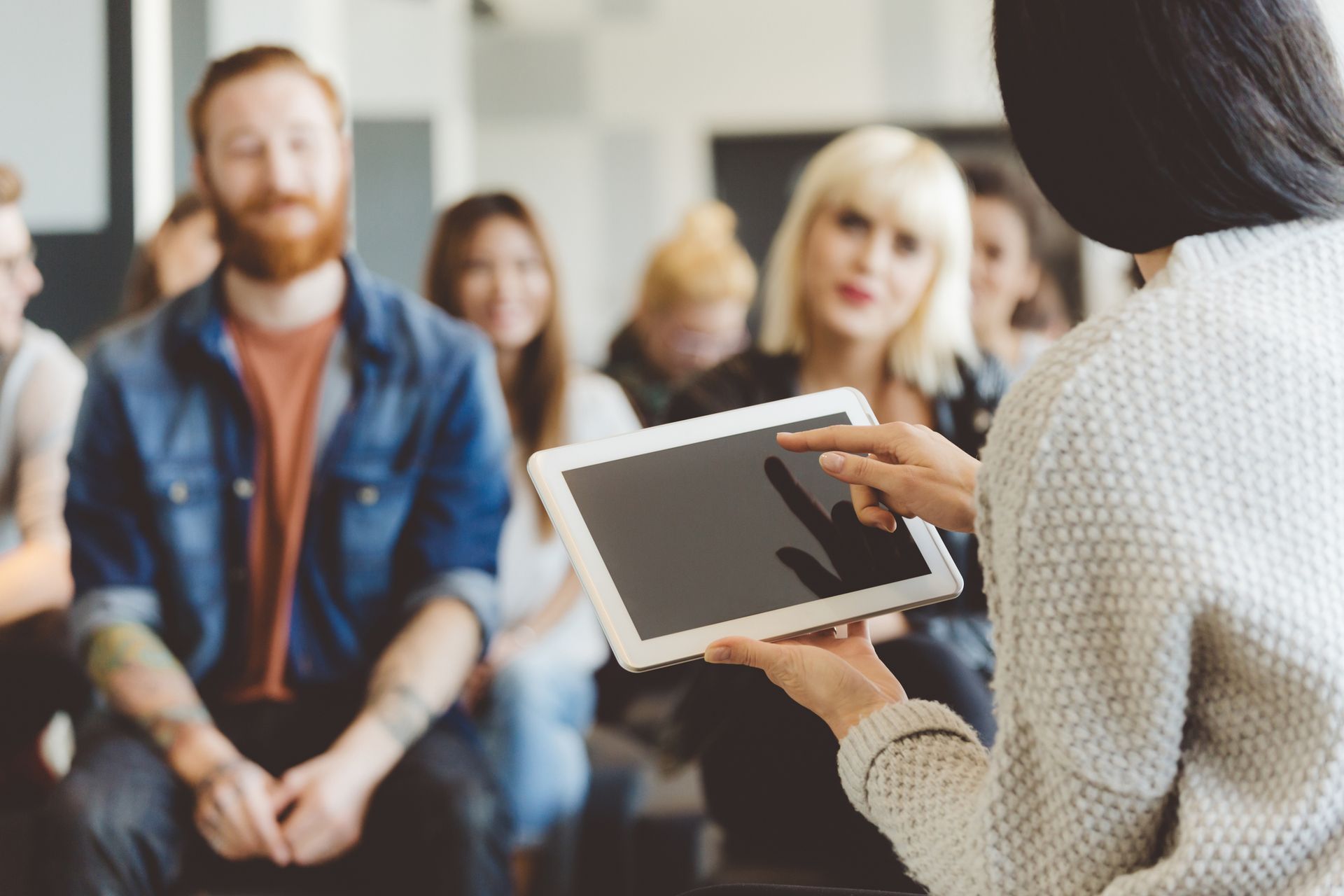 Woman presenting on a tablet to a diverse group seated indoors.