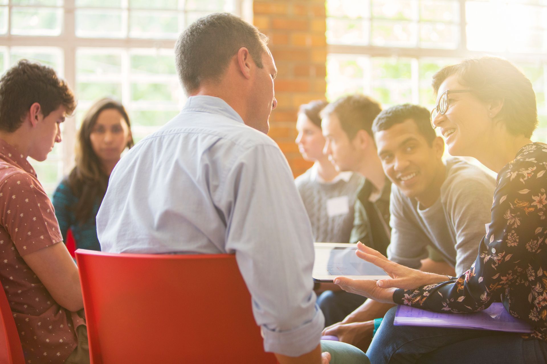 Group therapy session: People in a circle, some talking, in a room with a window, sunny.