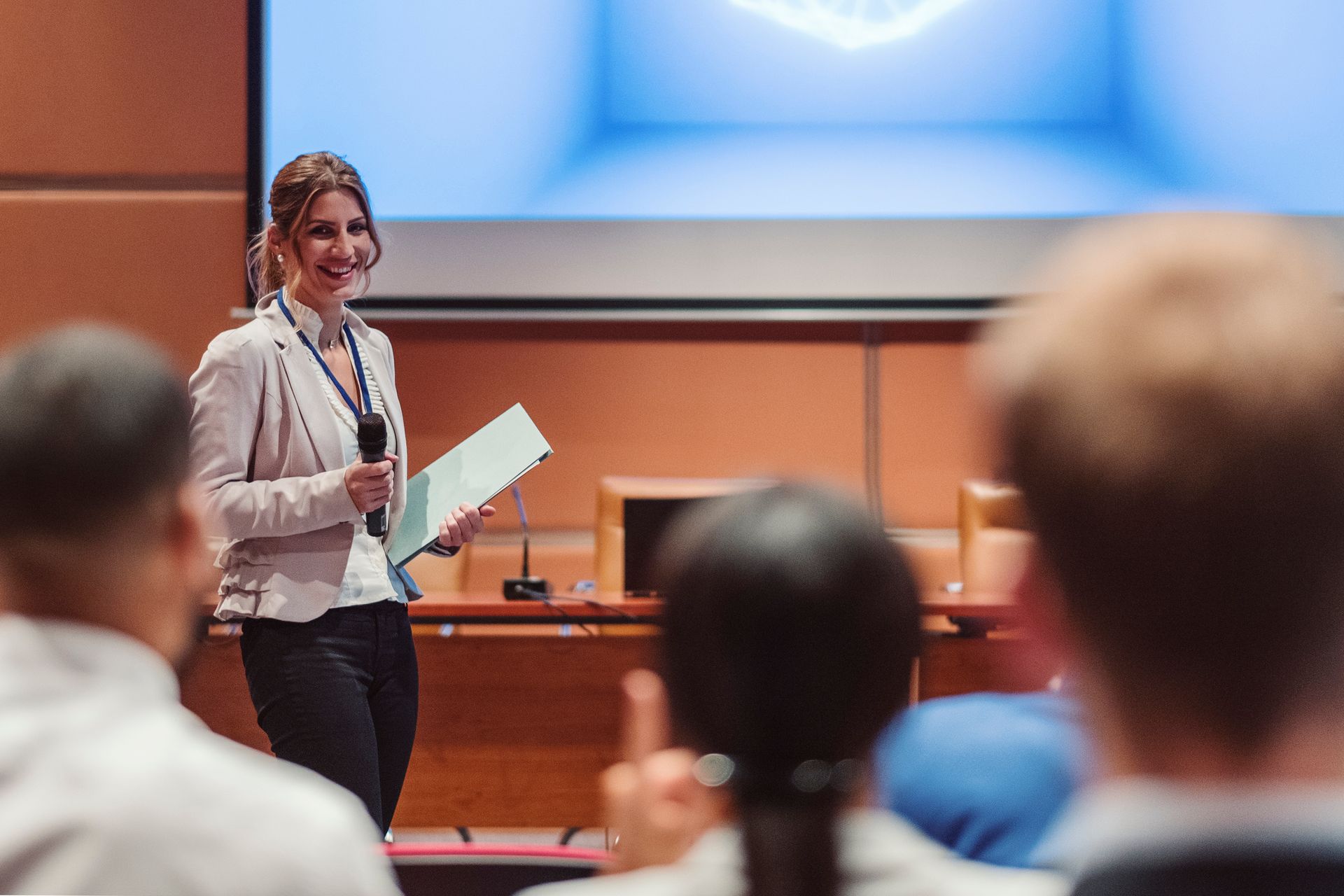 Woman speaking at a conference, holding a microphone and papers, with a screen in the background.
