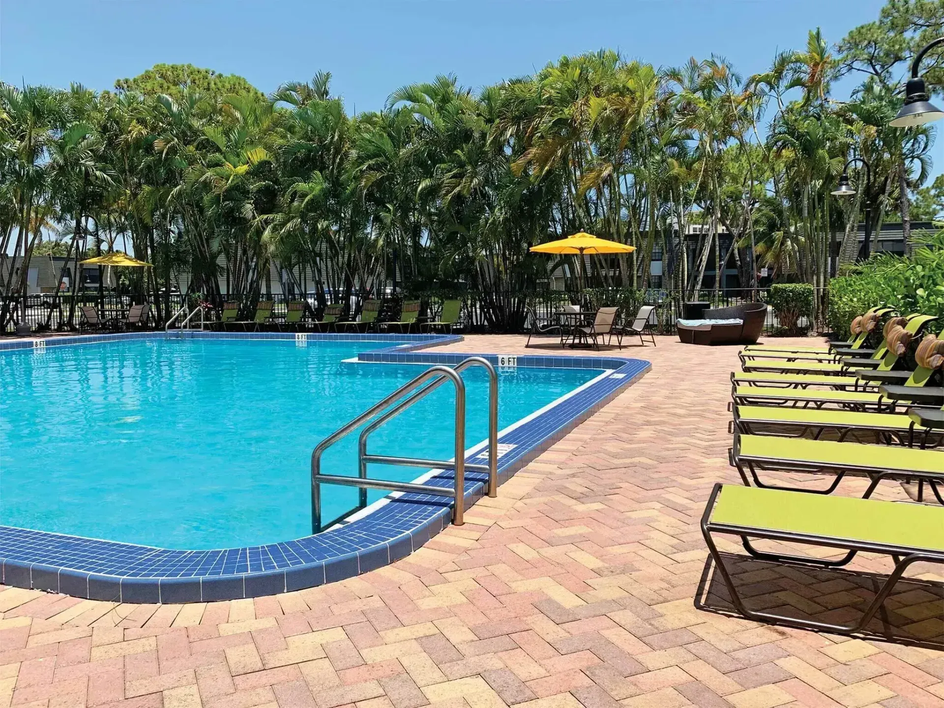 Swimming pool with blue water, surrounded by brick patio, lounge chairs, and palm trees.
