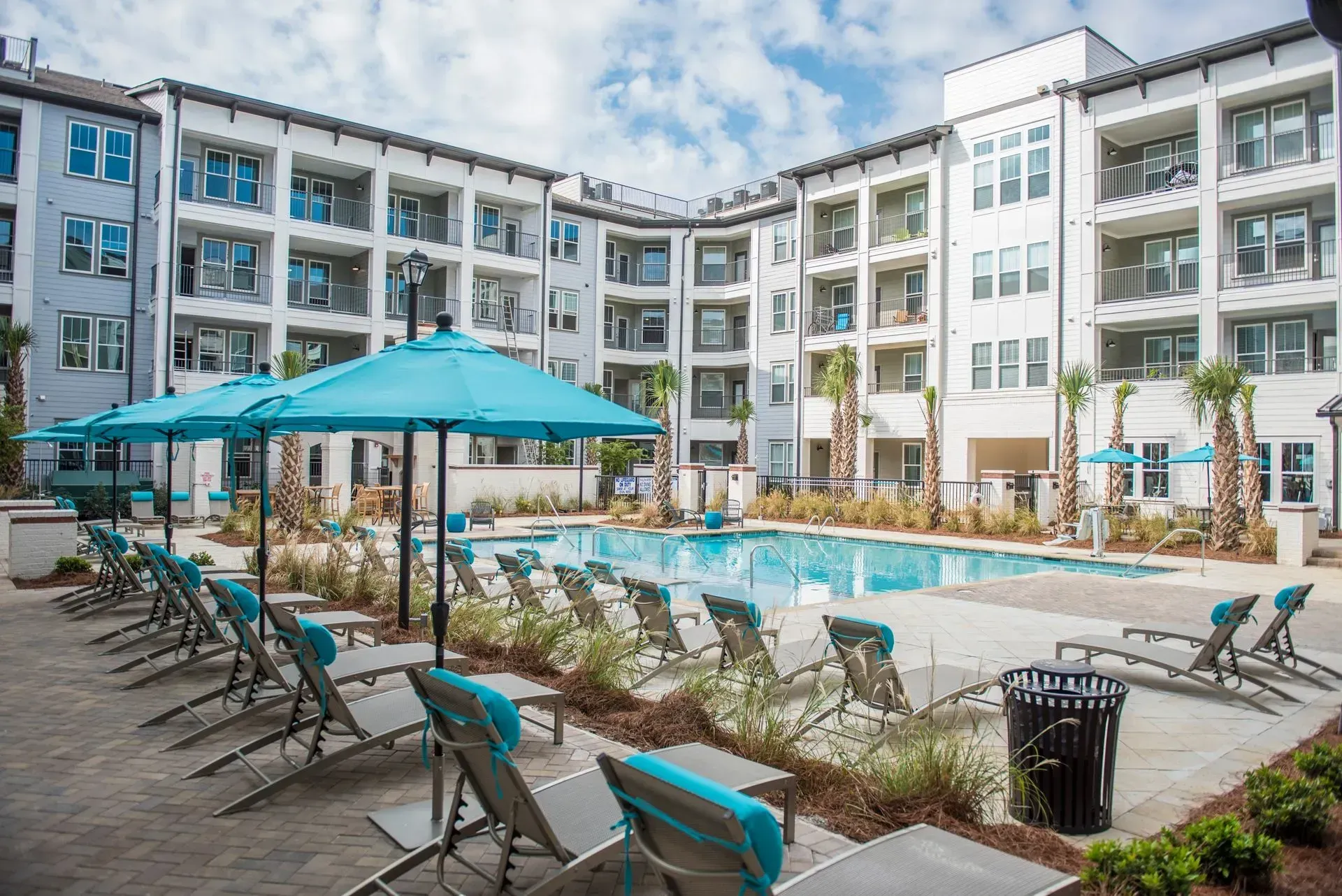 Pool area with turquoise umbrellas and lounge chairs, surrounded by a multi-story white apartment building.