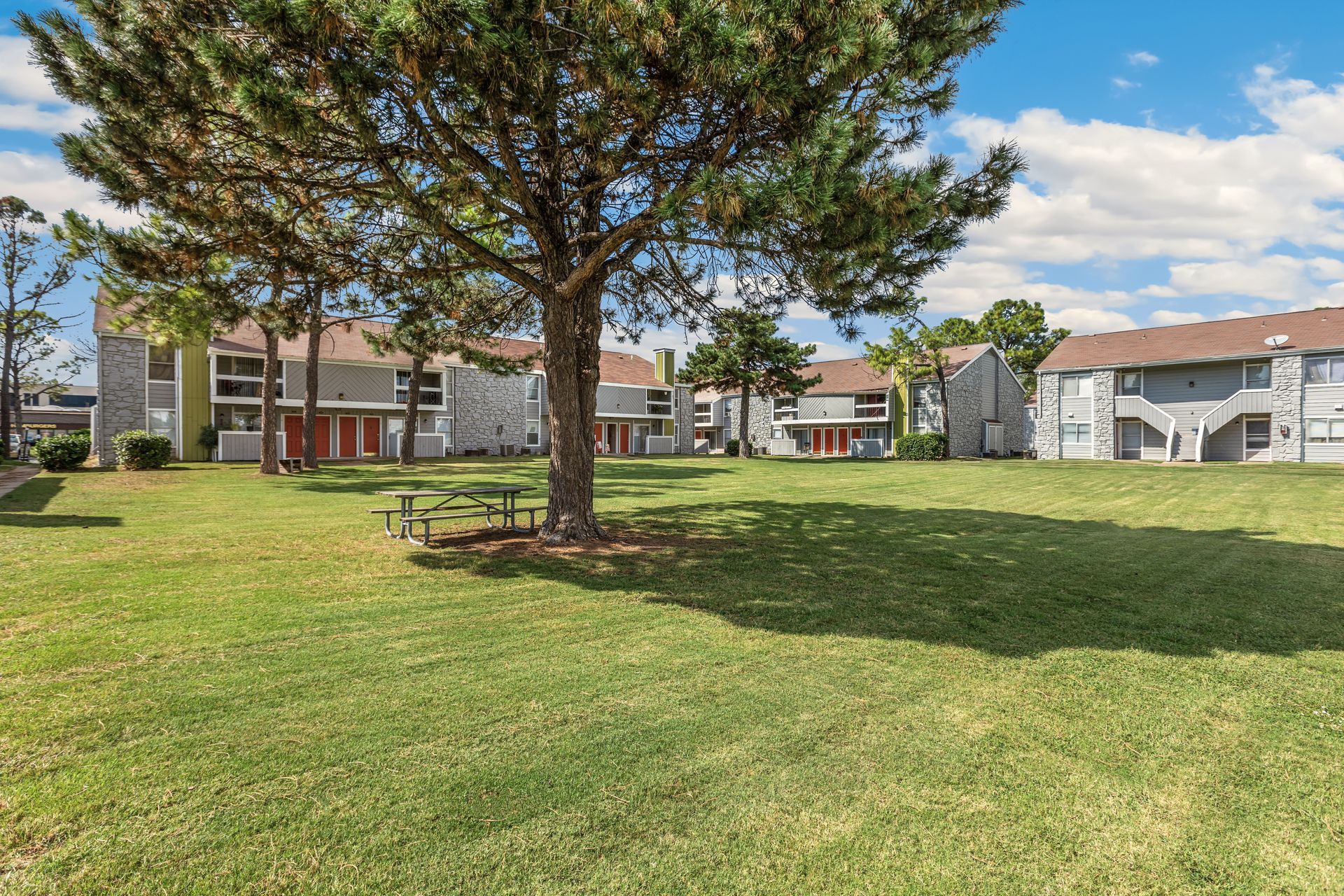 Grey buildings with white trim, green and orange accents. Lush green grass with trees, open space and blue skies.