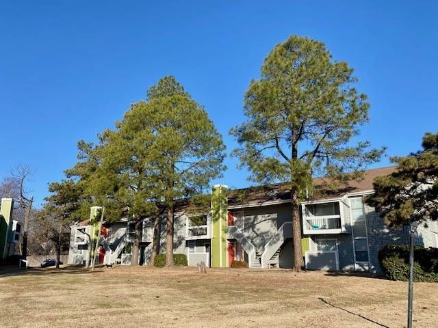 Apartment buildings with gray siding, green trim, and large trees against a clear blue sky.