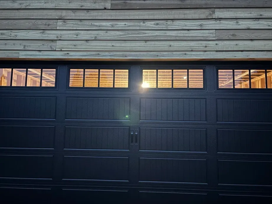 A modern black garage door with rectangular windows, positioned beneath horizontal wood siding.