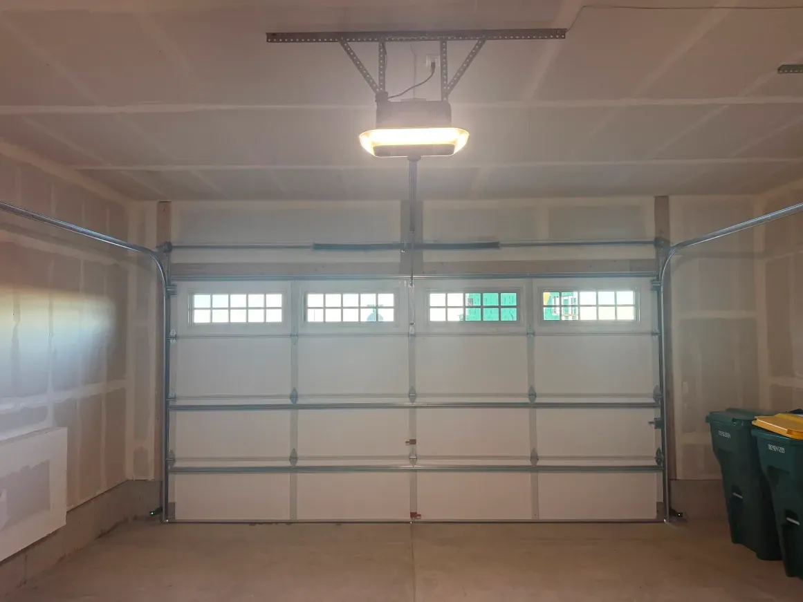 A white, multi-paneled garage door with windows, centered under a ceiling-mounted garage door opener in a finished garage.