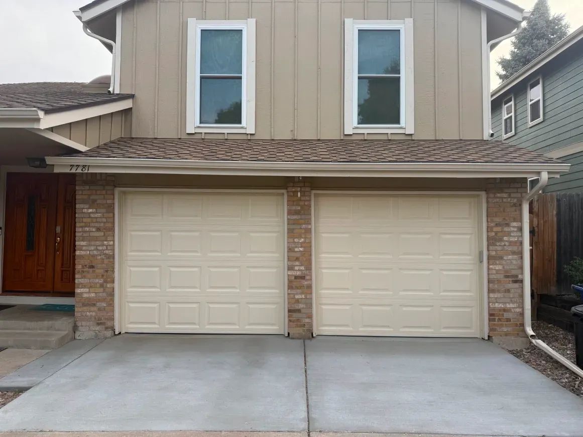 Two-story house exterior featuring tan siding, a brown brick facade, two beige garage doors, and a wood front door.